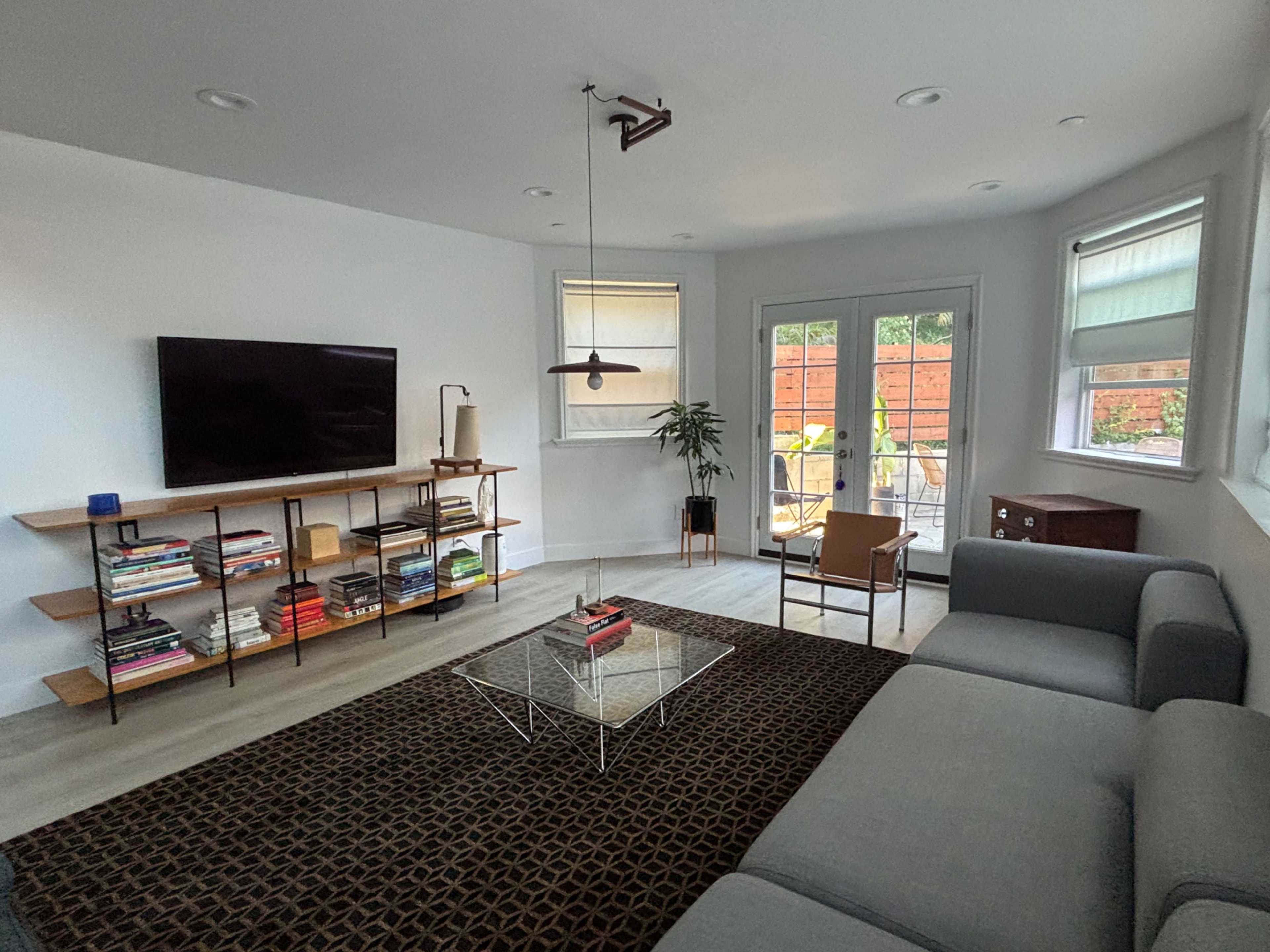 The image shows a modern living room with a sectional sofa, a glass coffee table, a wooden shelving unit with books, and large windows leading to an outdoor patio.