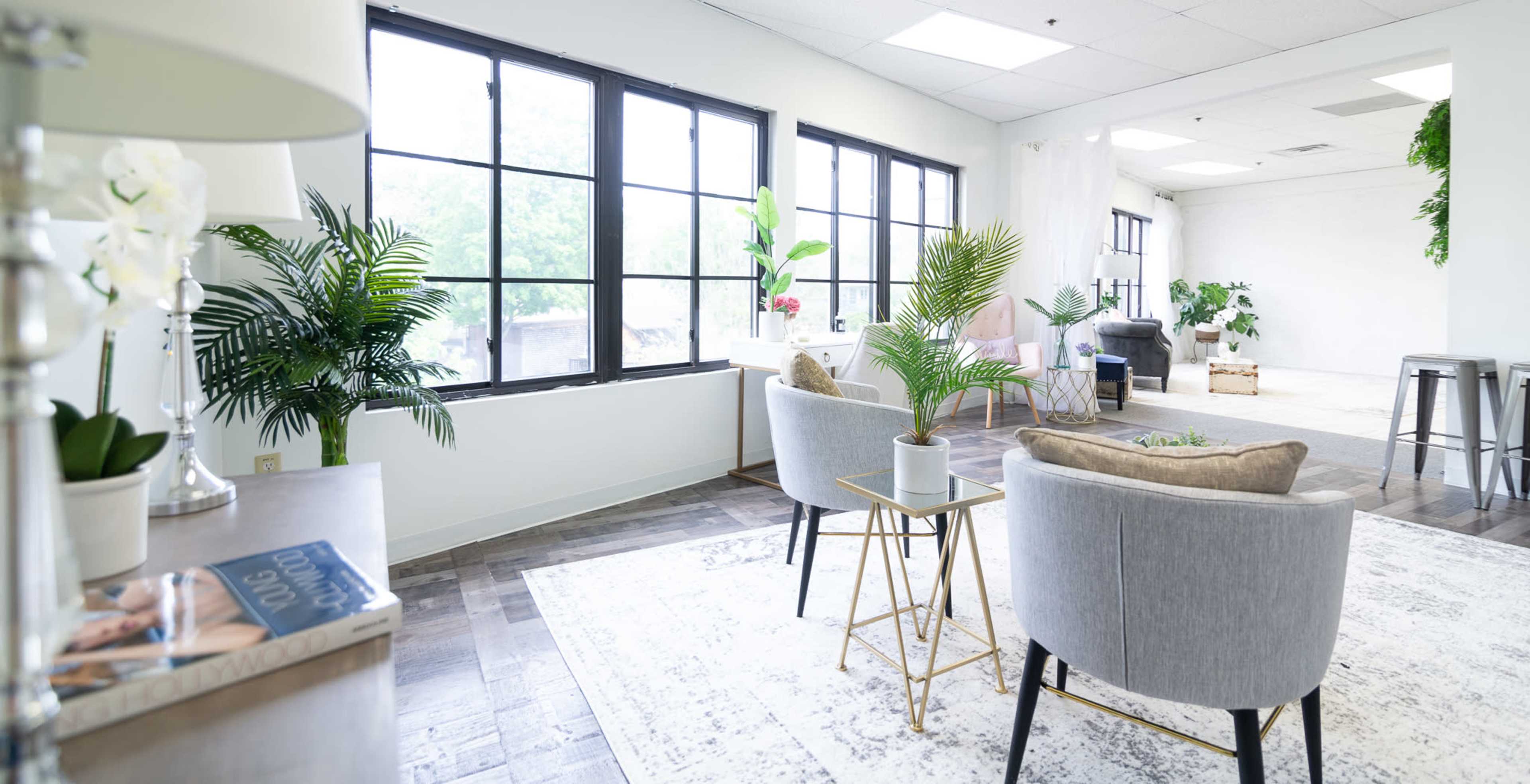 A modern office interior with large windows, grey chairs, a small table with a plant, and decorative greenery.