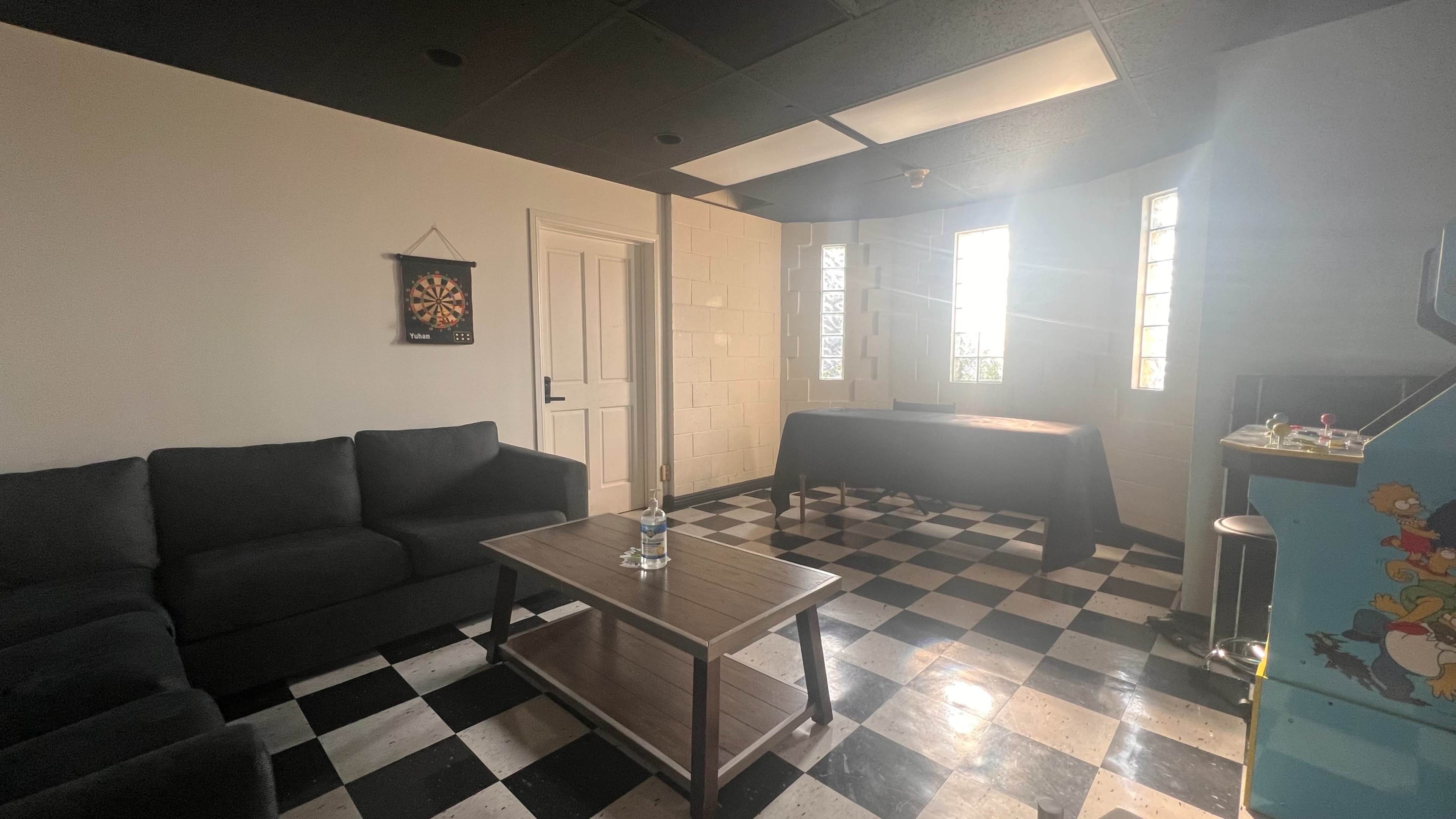 A room with a checkered floor features a dark sofa, a wooden table, and a window allowing light to enter.