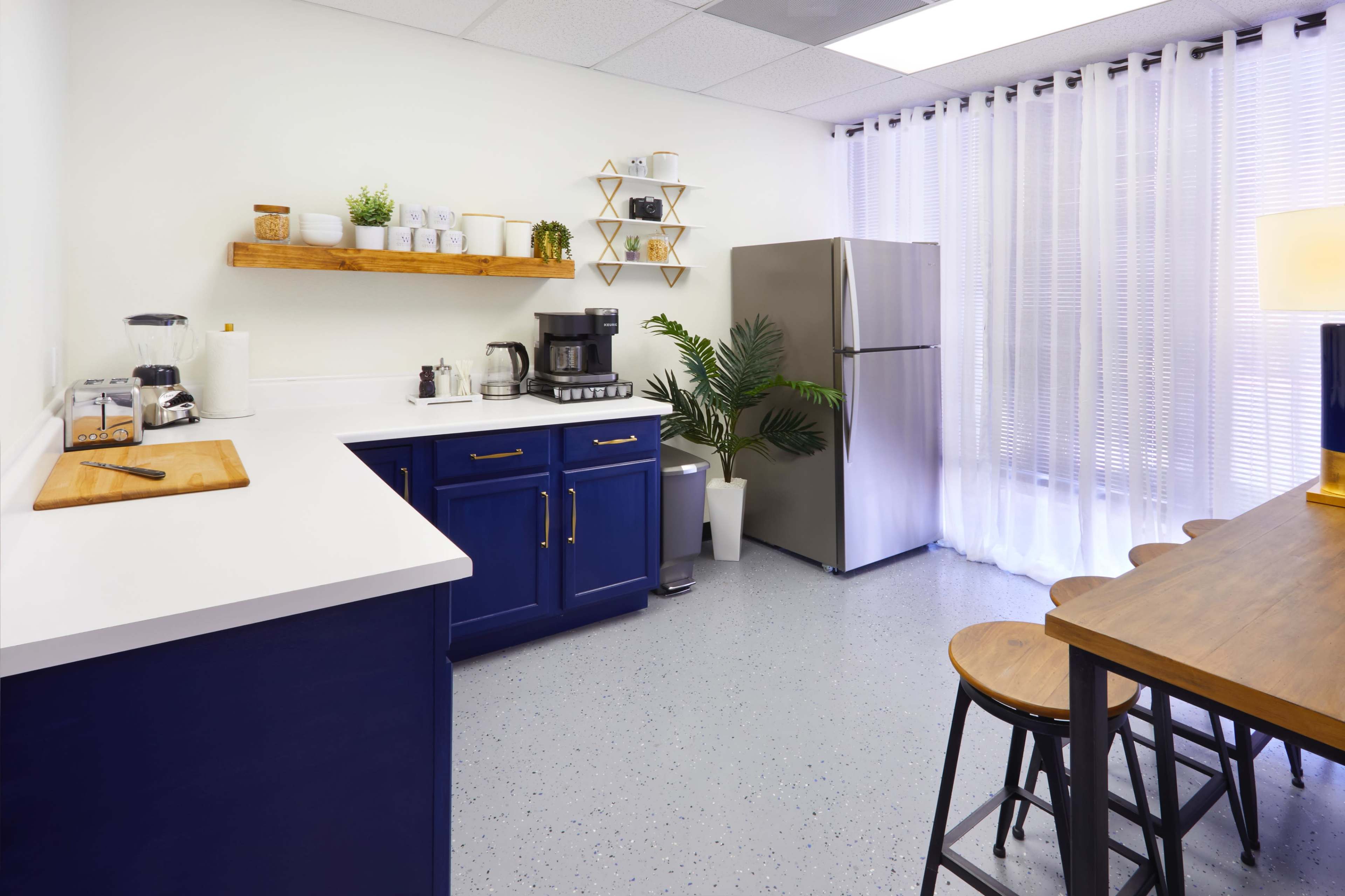 A modern kitchen features blue cabinets, a stainless steel refrigerator, a wood shelf with plants, and a dining area with wooden stools.