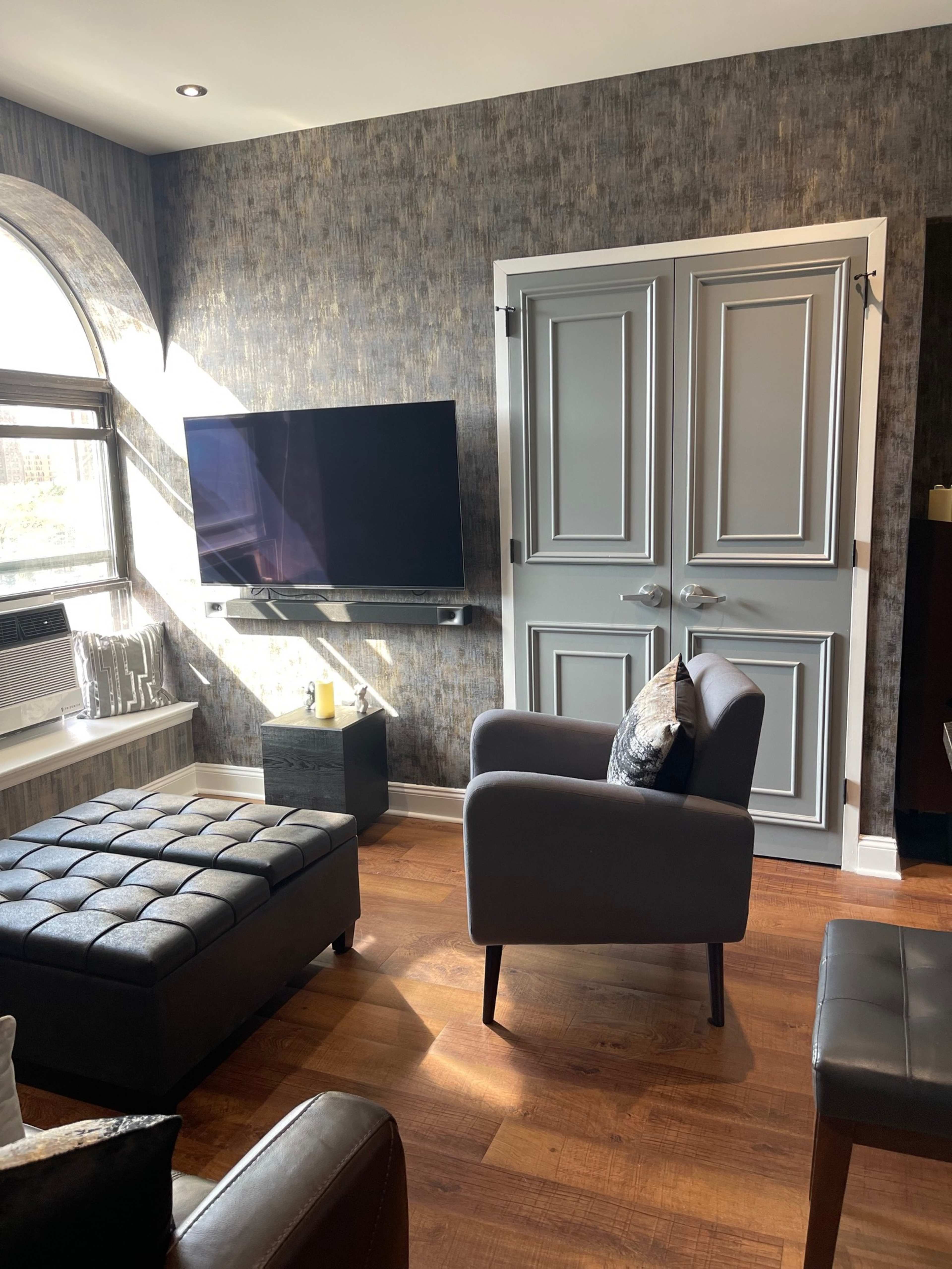 A modern living room features a flat-screen TV mounted on the wall, a dark tufted ottoman, and a pair of closed double doors near a large window.