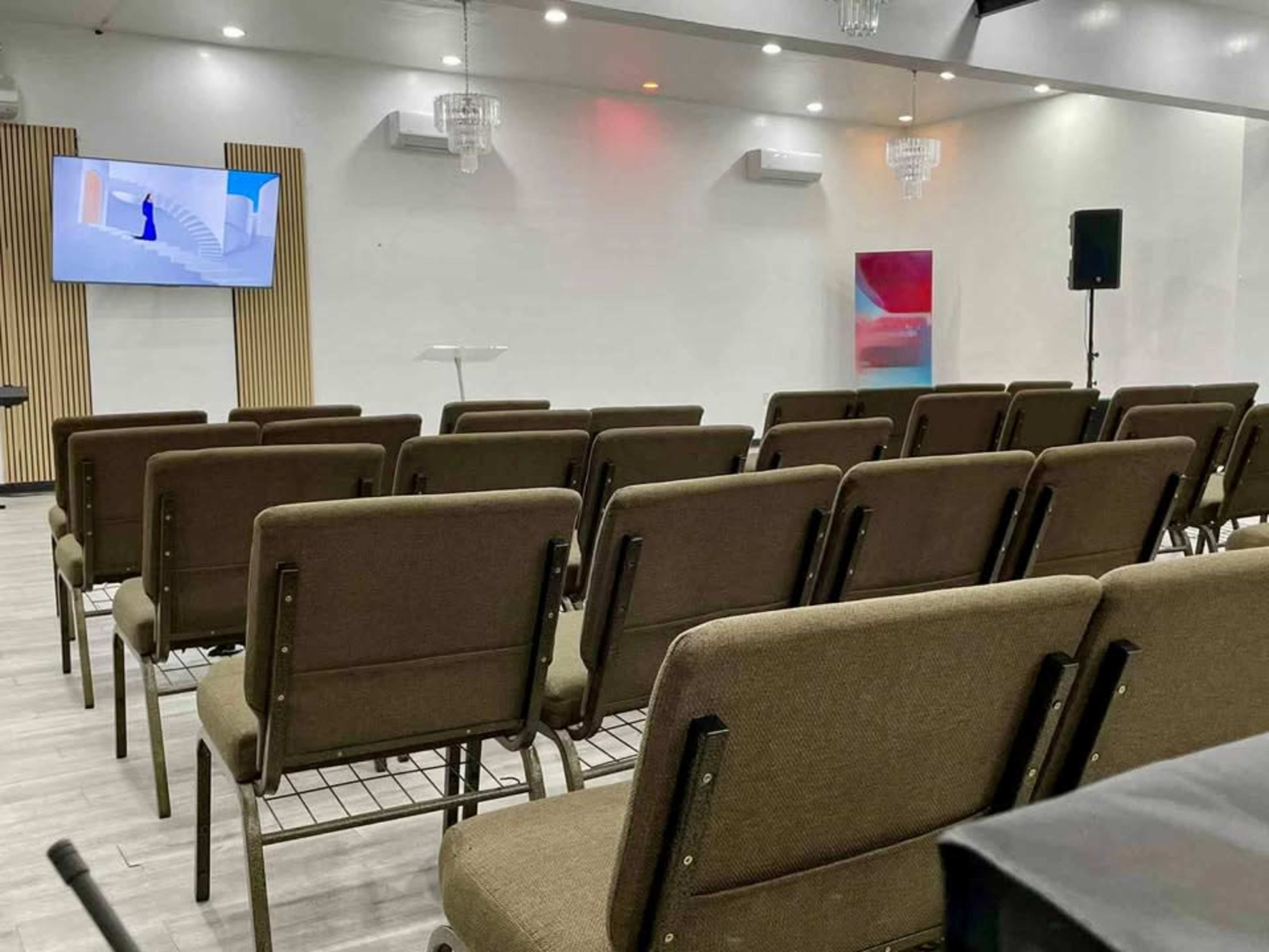 The image shows a spacious event room with chairs arranged in rows facing a screen displaying a presentation, illuminated by overhead lights.