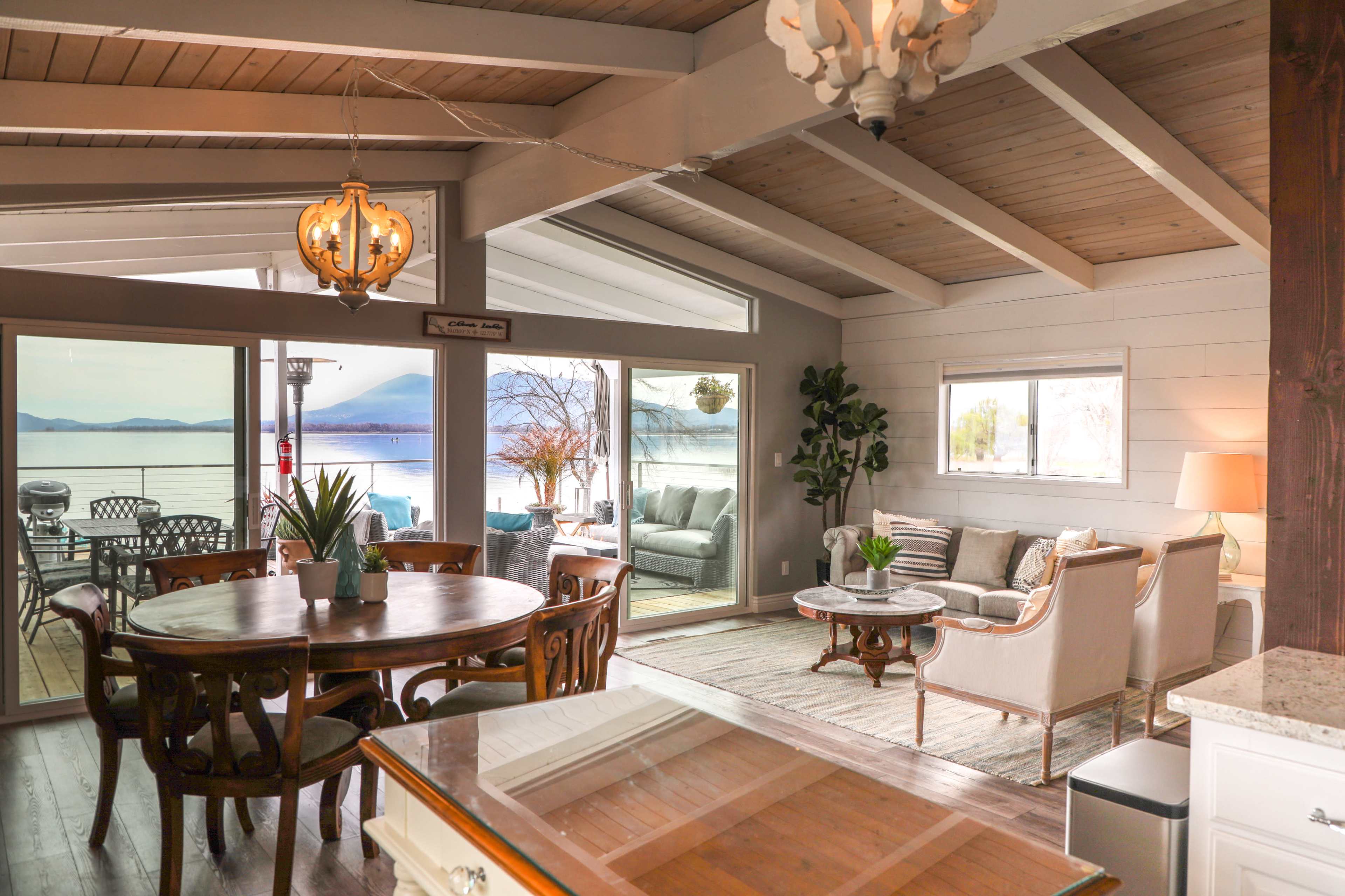 An open-concept living and dining area with large windows showcasing a water view, featuring a round dining table, comfortable seating, and wooden beams on the ceiling.