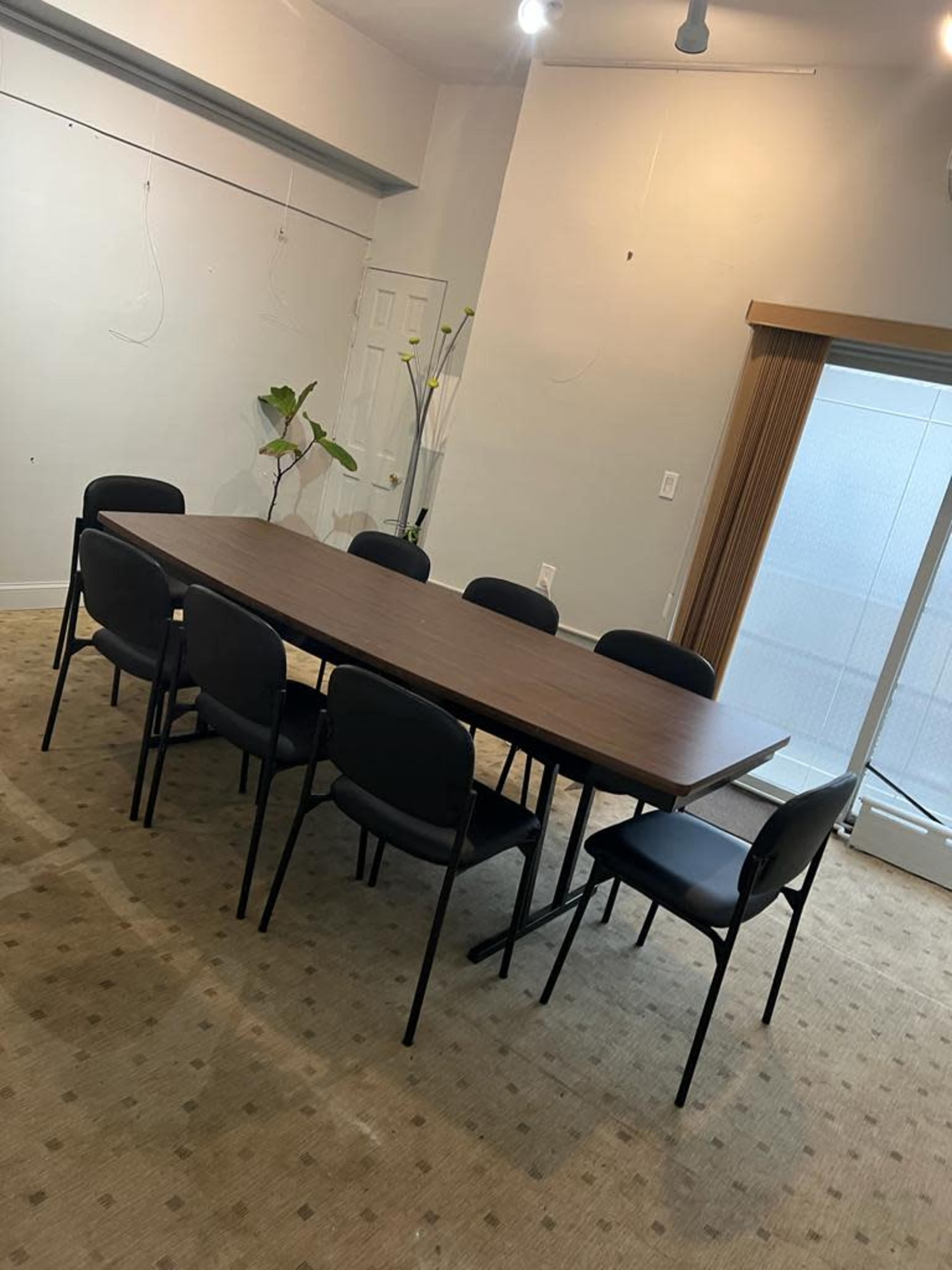 A rectangular wooden table surrounded by eight black chairs is set in a room with a sliding door and a plant in the corner.