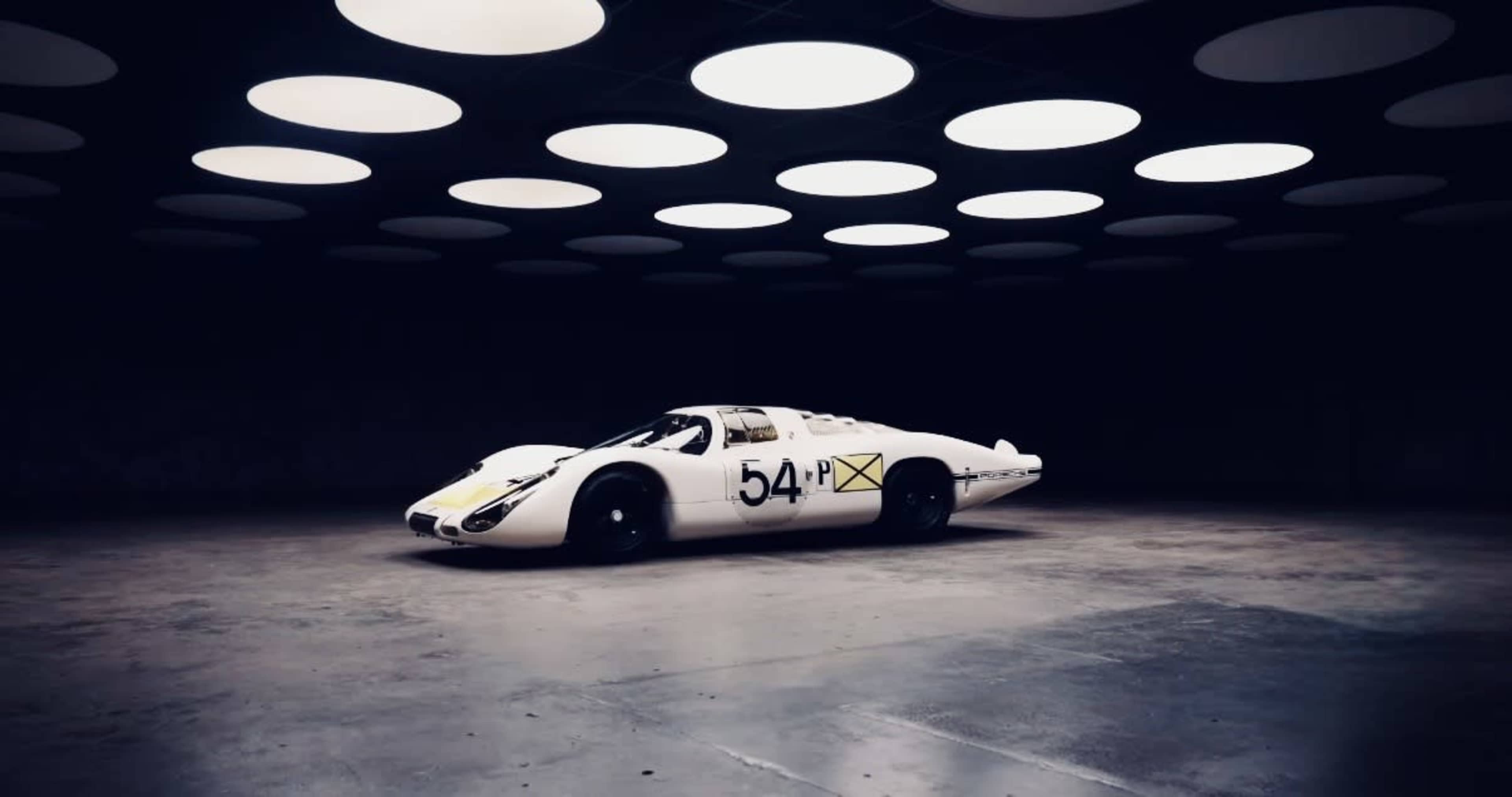A vintage race car with the number 54 painted on it is positioned in a dimly lit garage under circular ceiling lights.