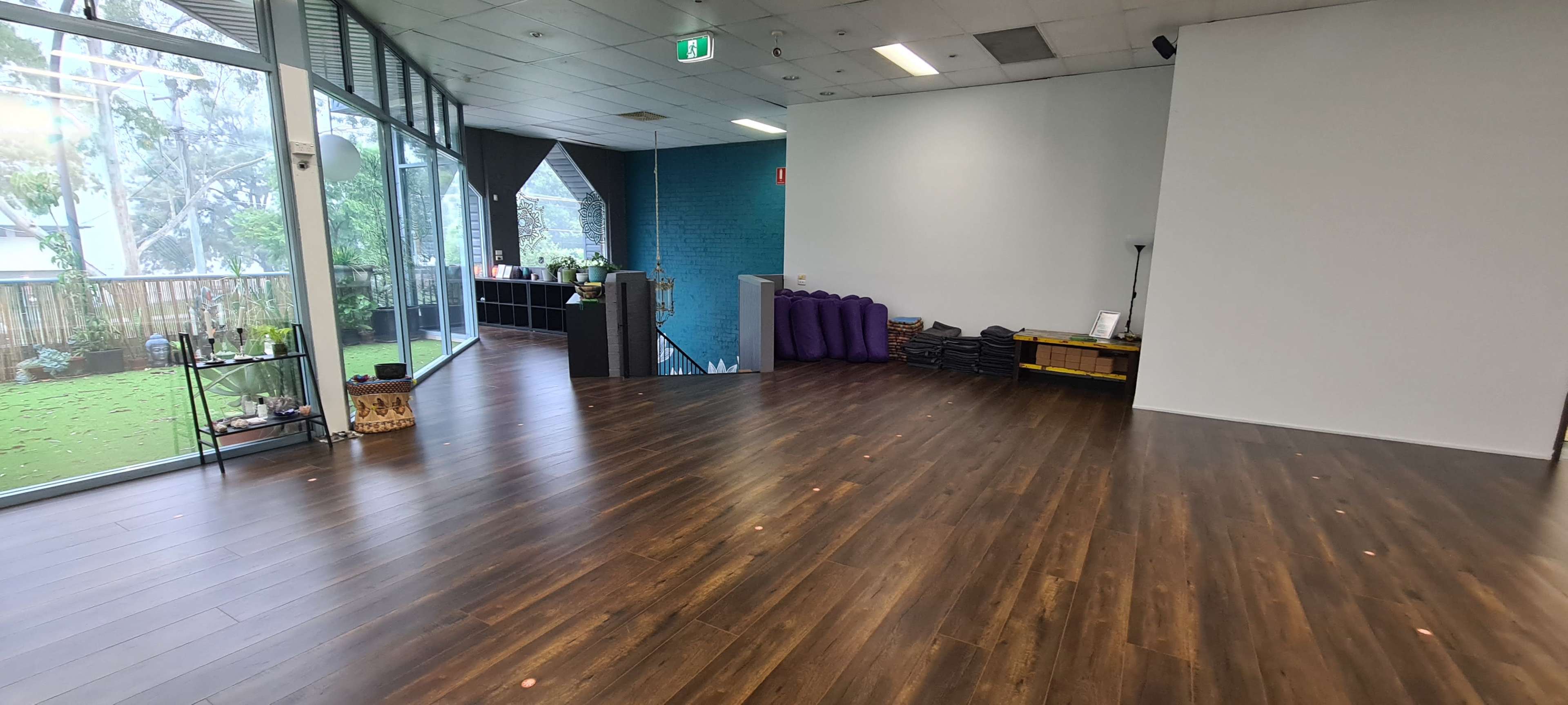 The image shows a spacious, empty room with wooden flooring, large windows revealing greenery outside, and minimal furnishings along one wall.