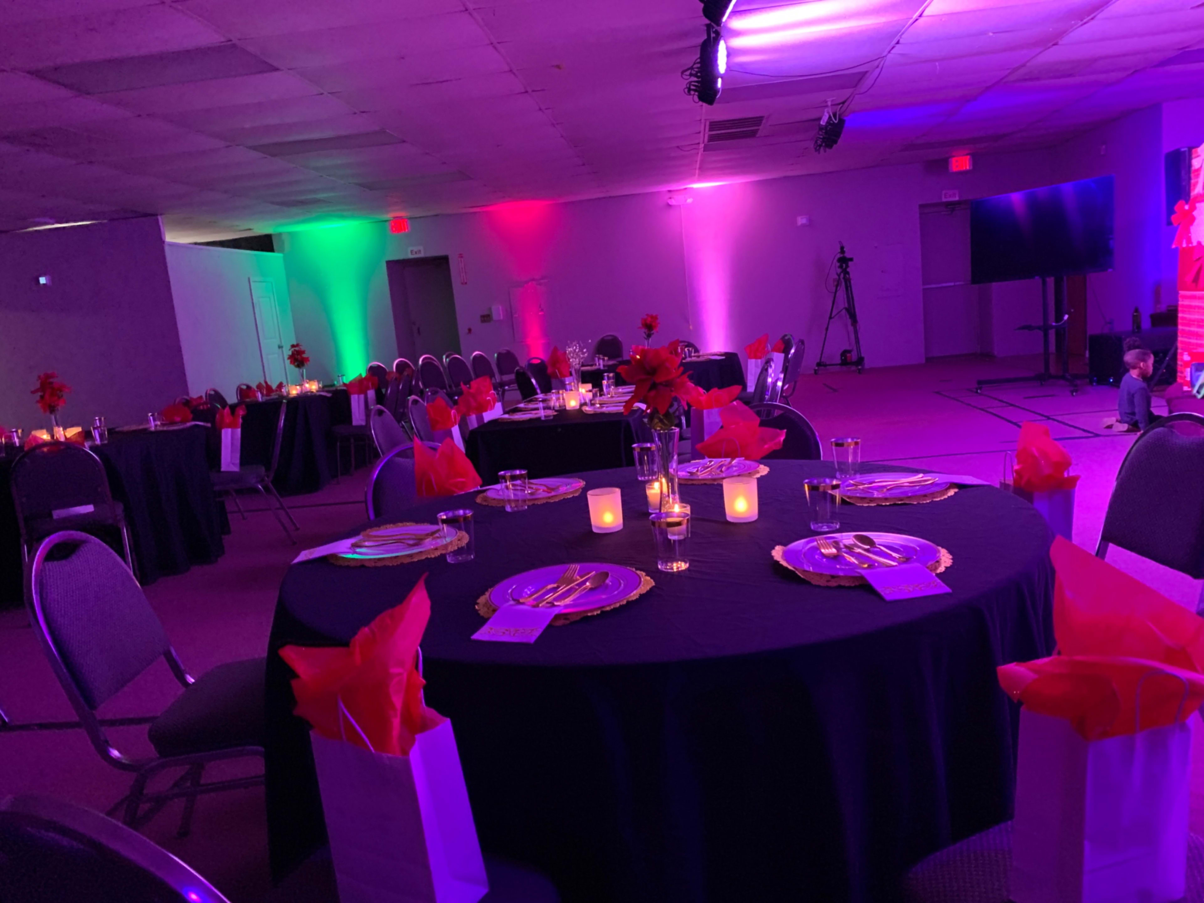 A banquet hall set for an event, featuring round tables adorned with black tablecloths, gold plates, candles, and gift bags, illuminated by colored lights.