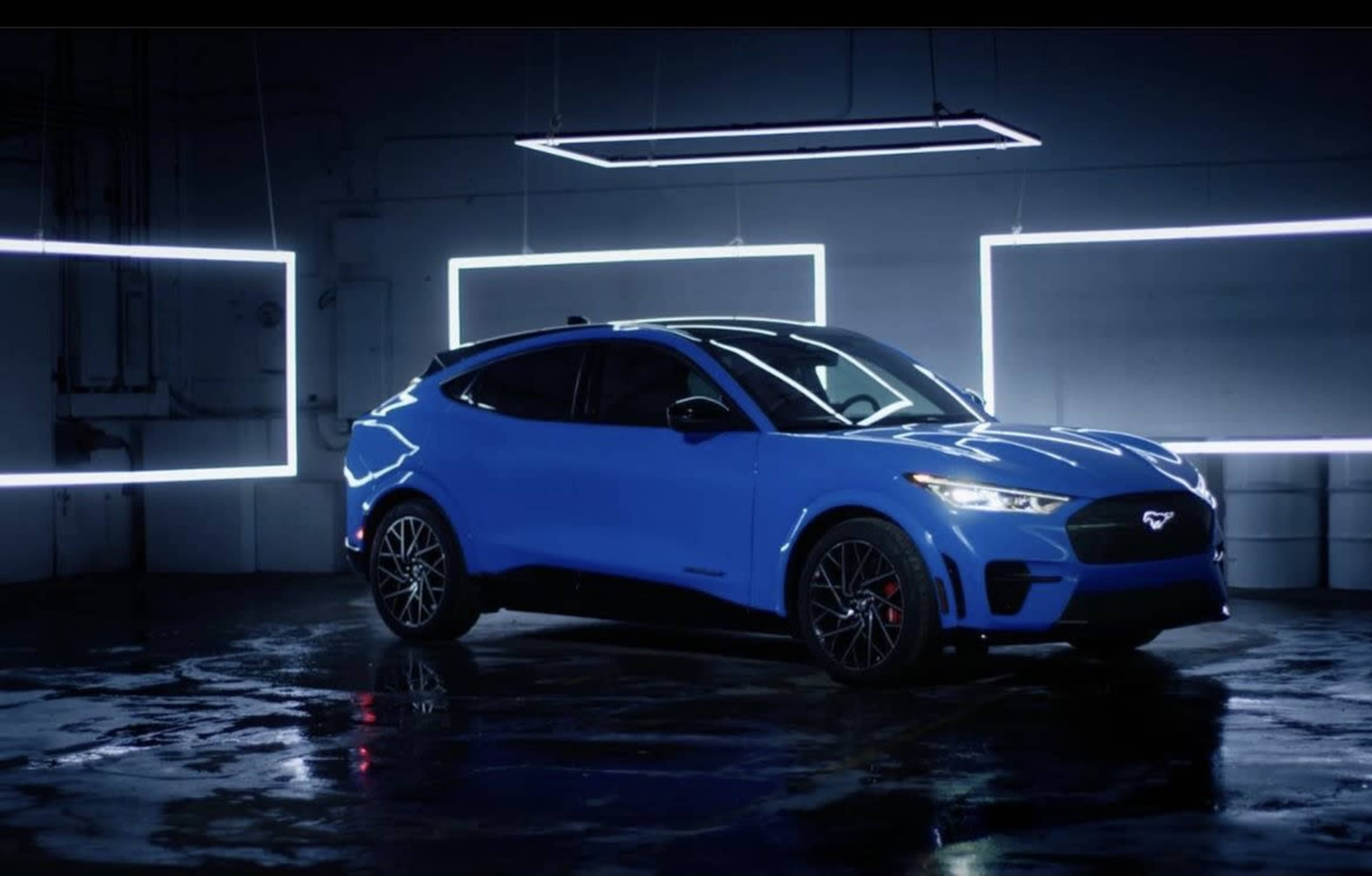 A blue electric car is displayed in a dark room illuminated by rectangular white light fixtures.