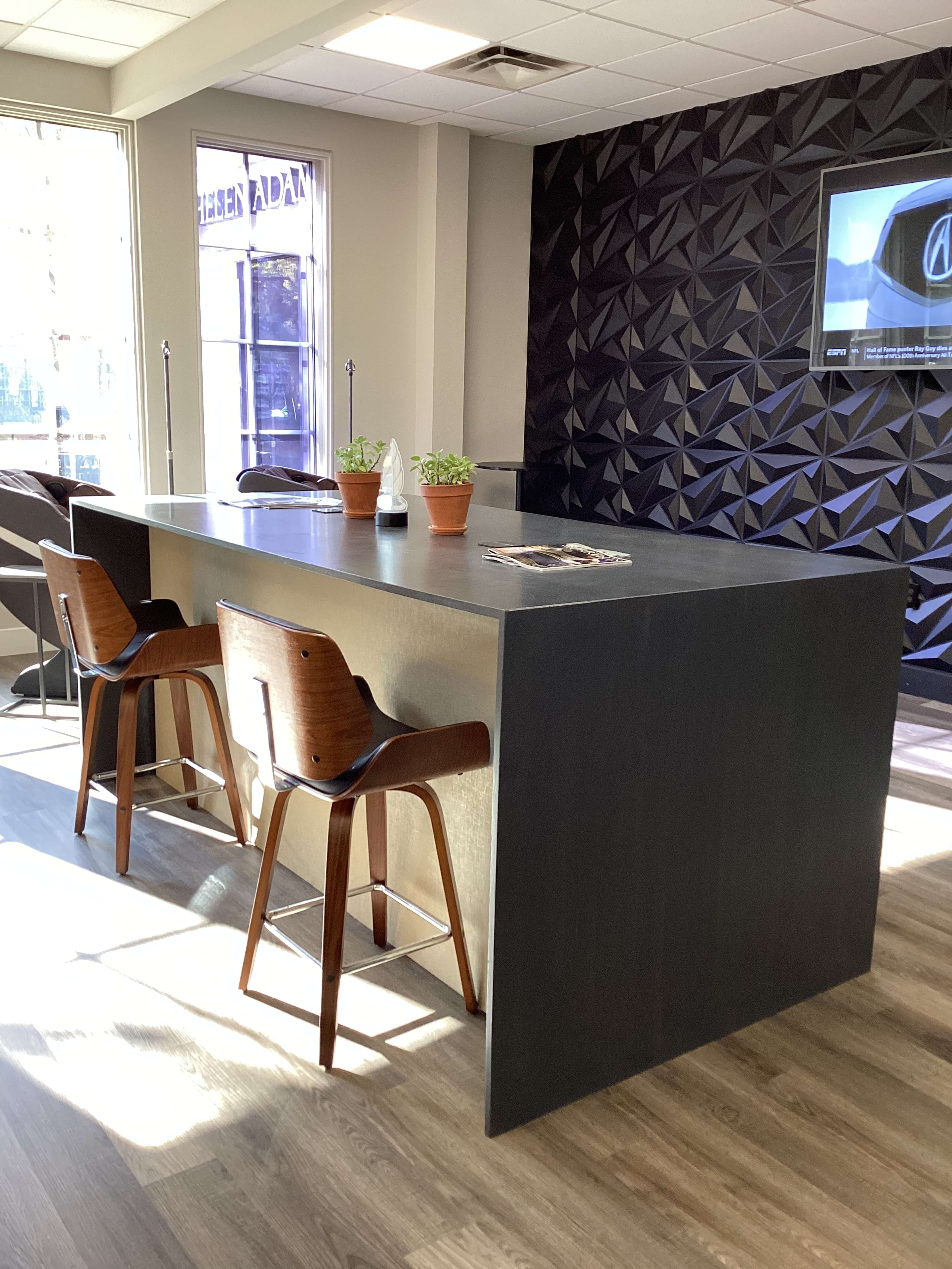 A modern reception area with a large dark counter and two wooden stools, illuminated by natural light from a nearby window.