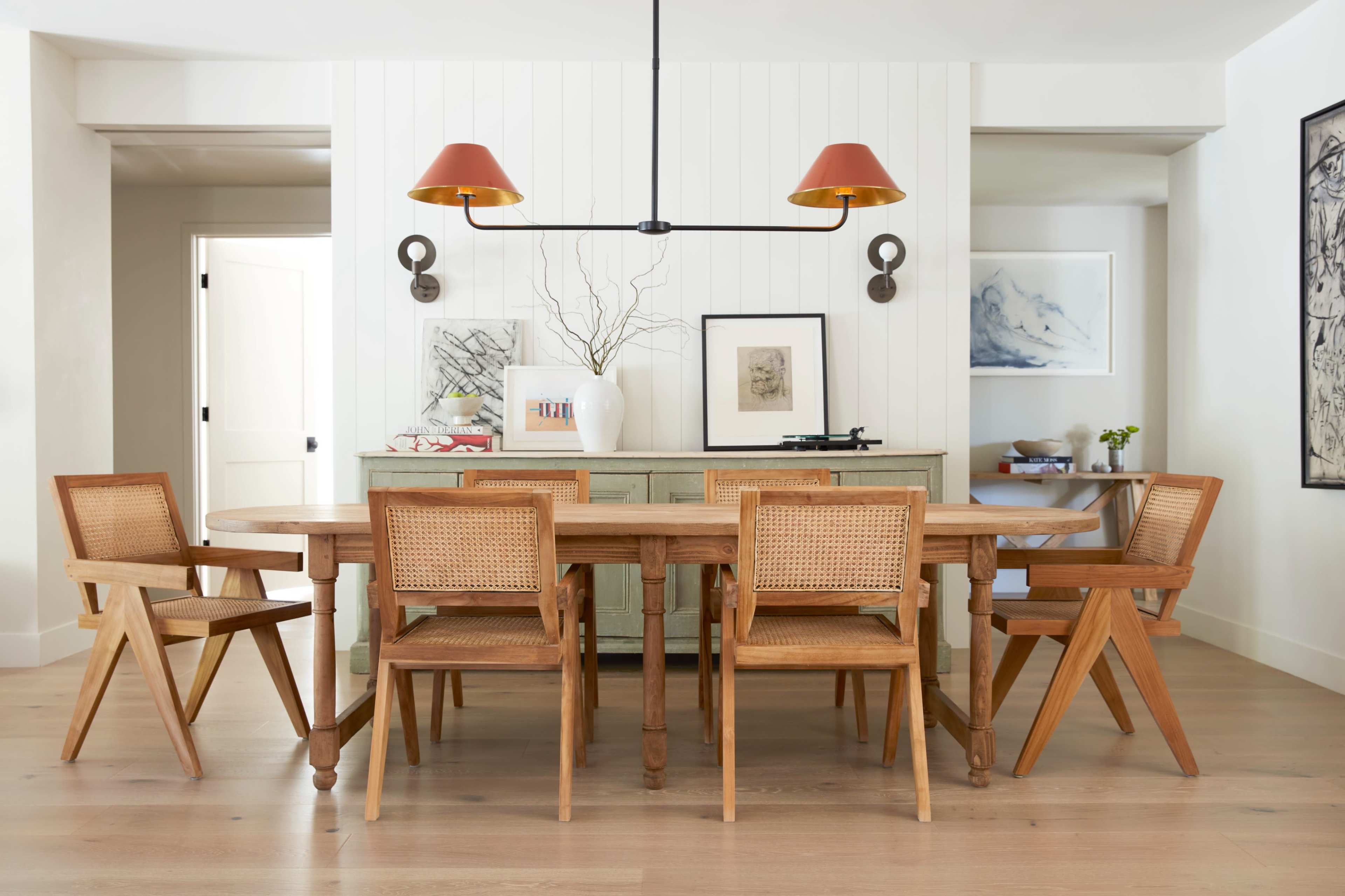 A wooden dining table is surrounded by six chairs in a well-lit room featuring a light-colored wall and pendant lighting.