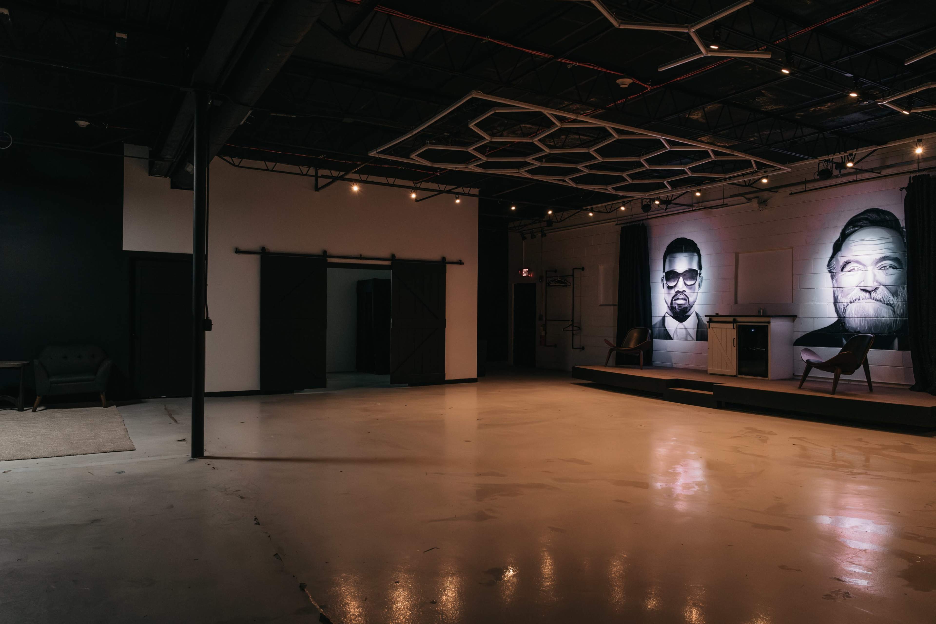 The image shows a spacious, dimly lit gallery with large portraits of two individuals on the wall and a raised platform at the front.