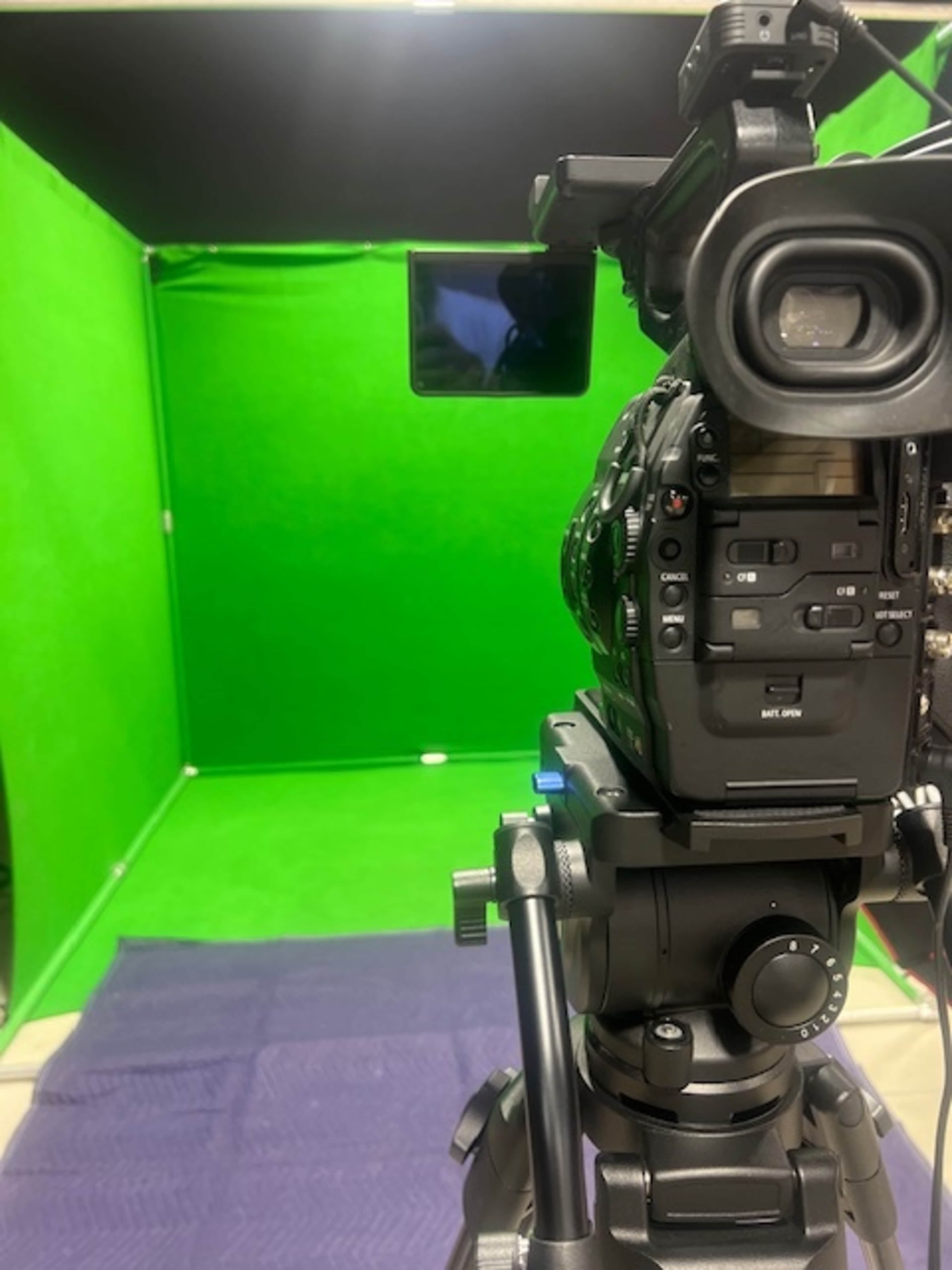 A video camera is set up on a tripod facing a green screen background in a studio environment.
