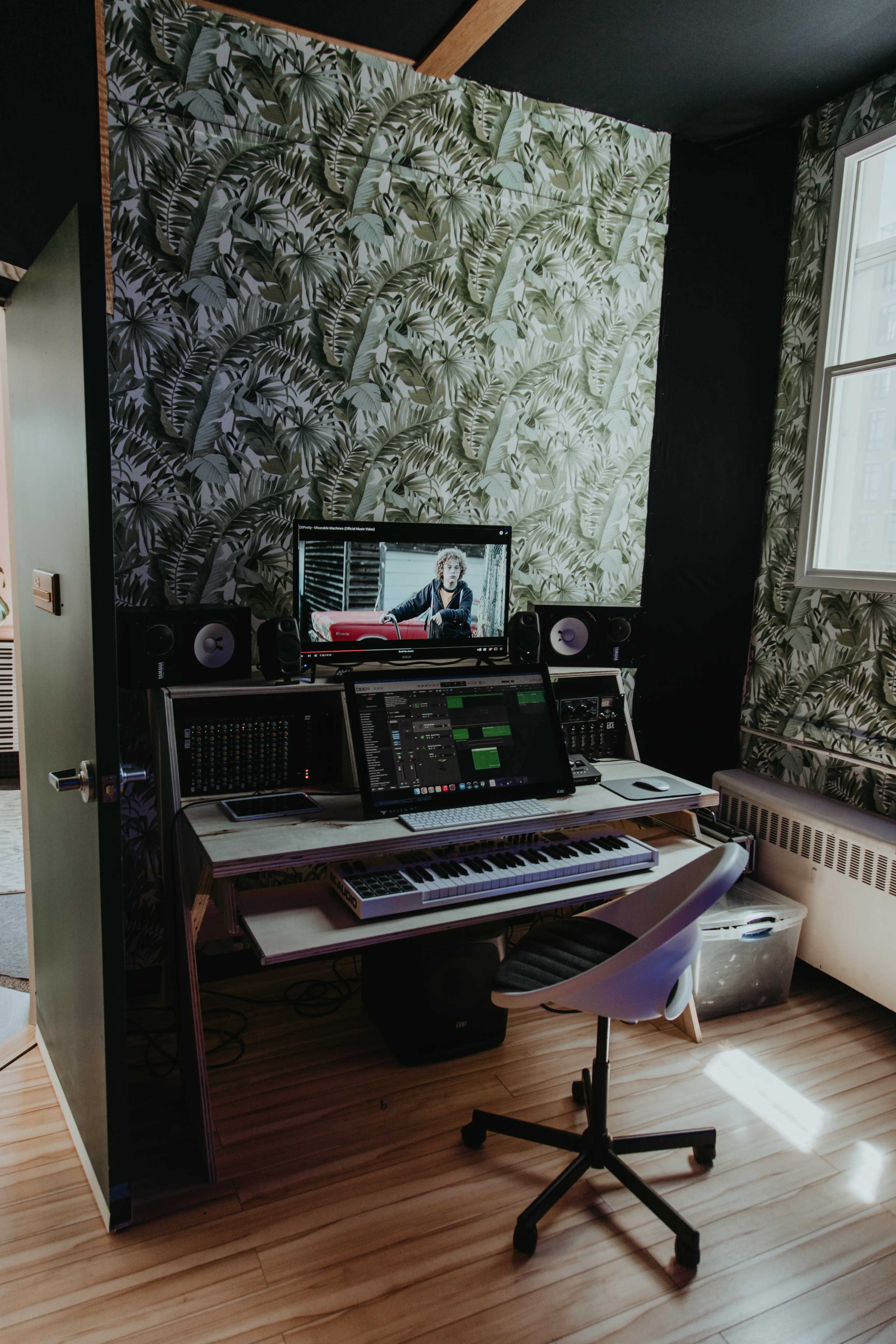 A music production workspace features a computer monitor with a video, a keyboard, and audio equipment against a backdrop of leafy wallpaper.