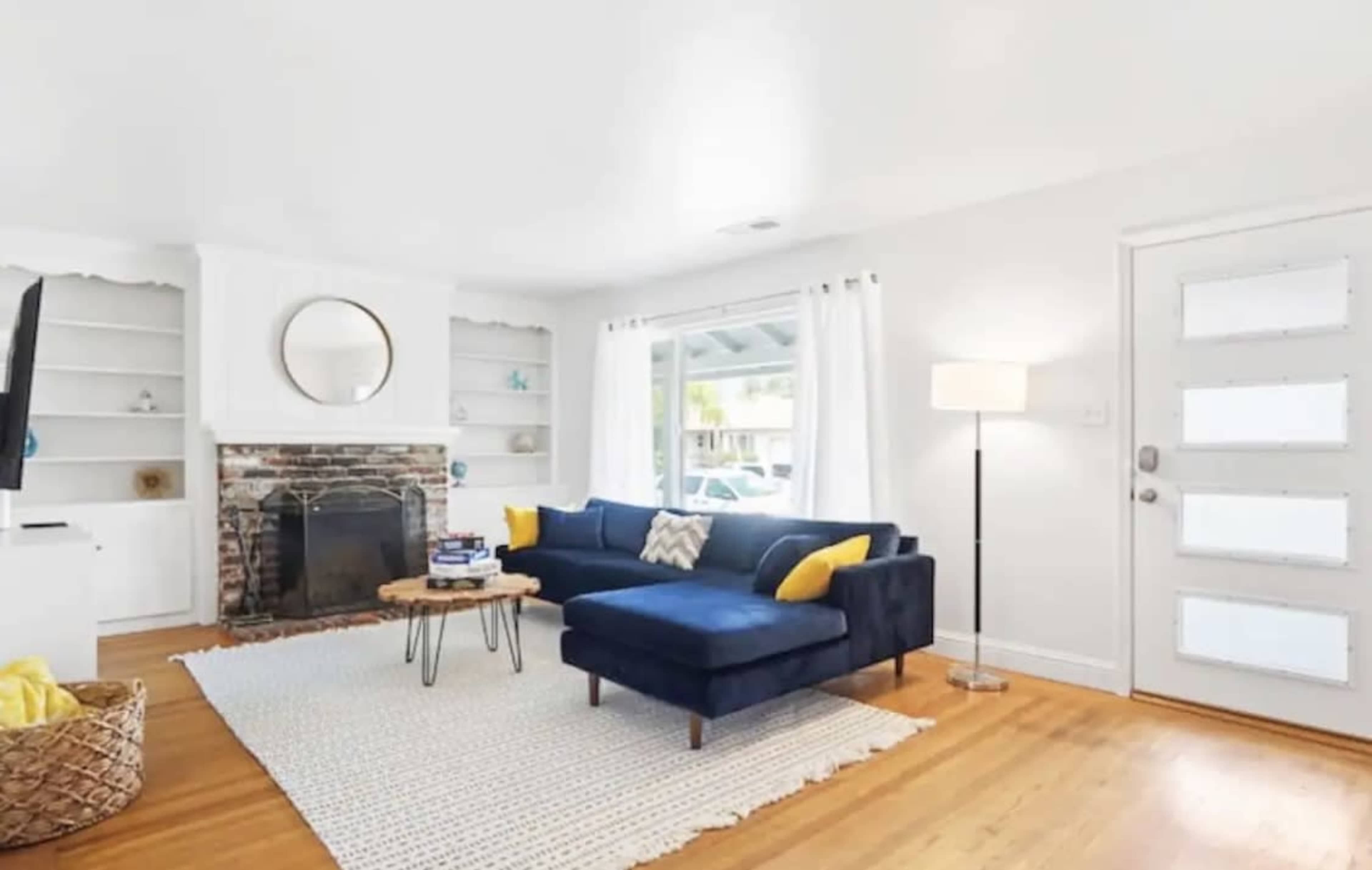 A living room features a blue couch with yellow cushions, a brick fireplace, a round mirror, and large windows, alongside a lamp and a woven basket.
