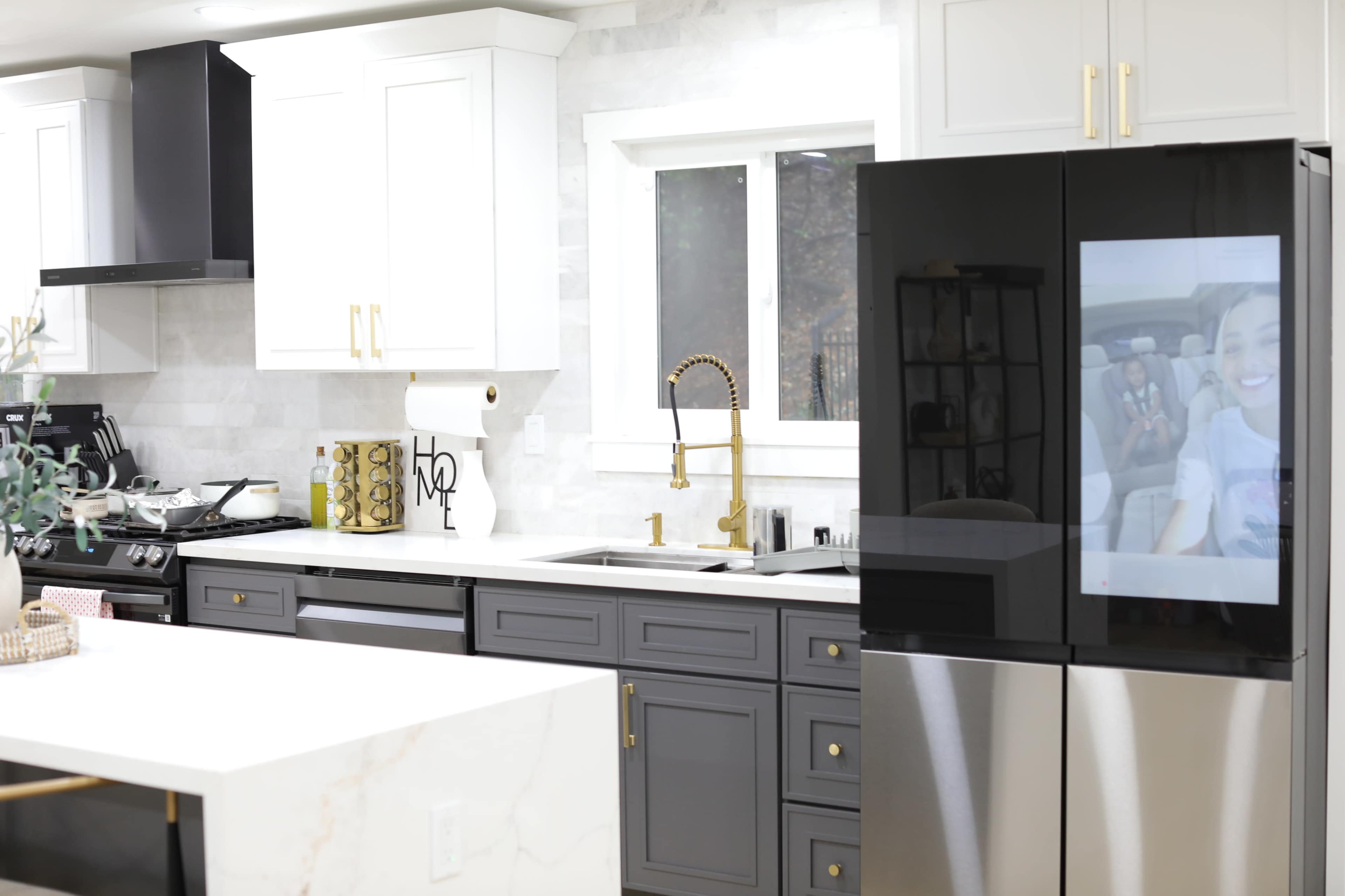 The kitchen features a combination of white and dark gray cabinetry, modern appliances, and a large refrigerator with a screen displaying a reflective image.