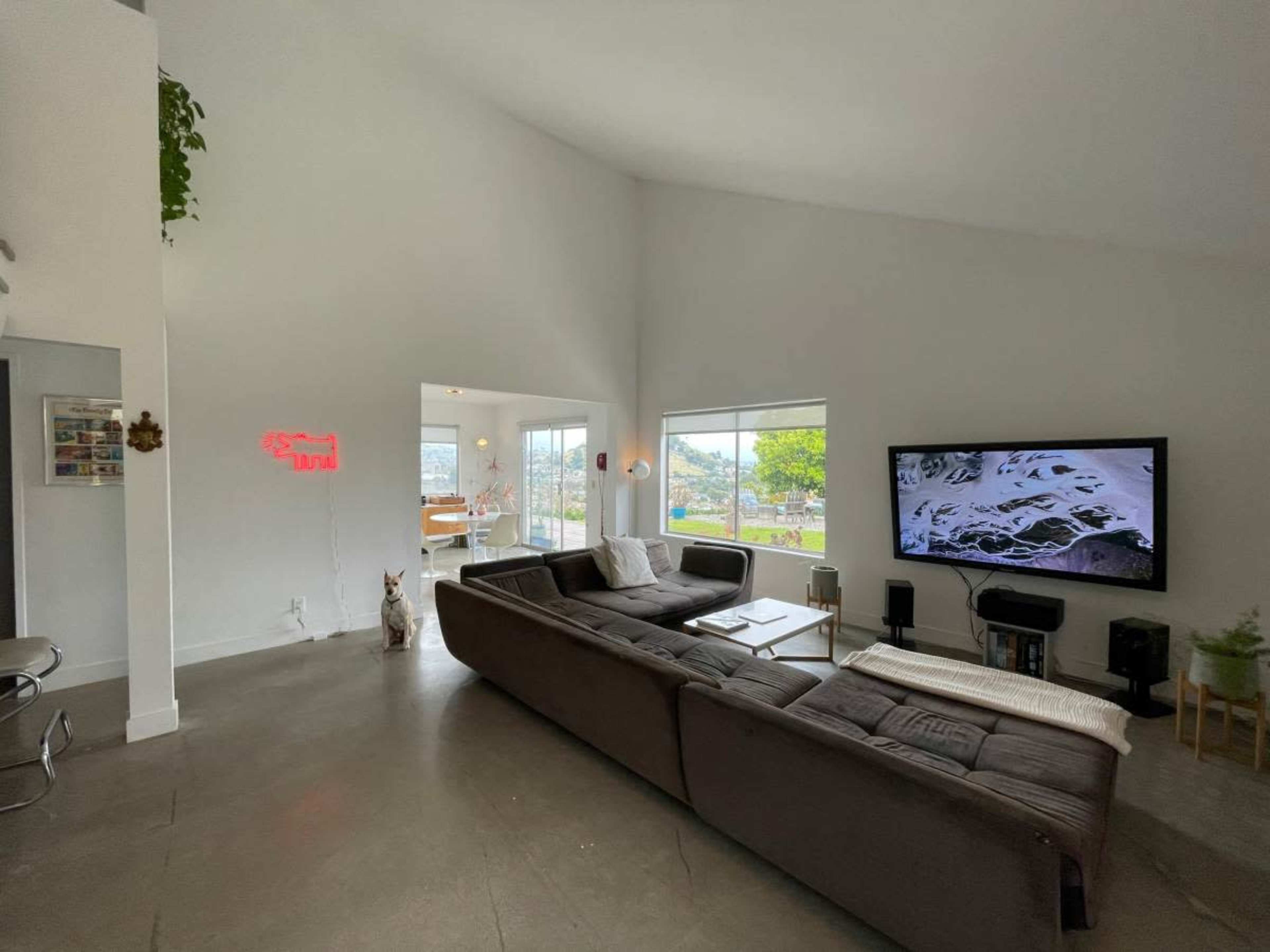 A modern living room features a large sectional sofa, a flat-screen TV, and windows that provide natural light and views of the outdoors.