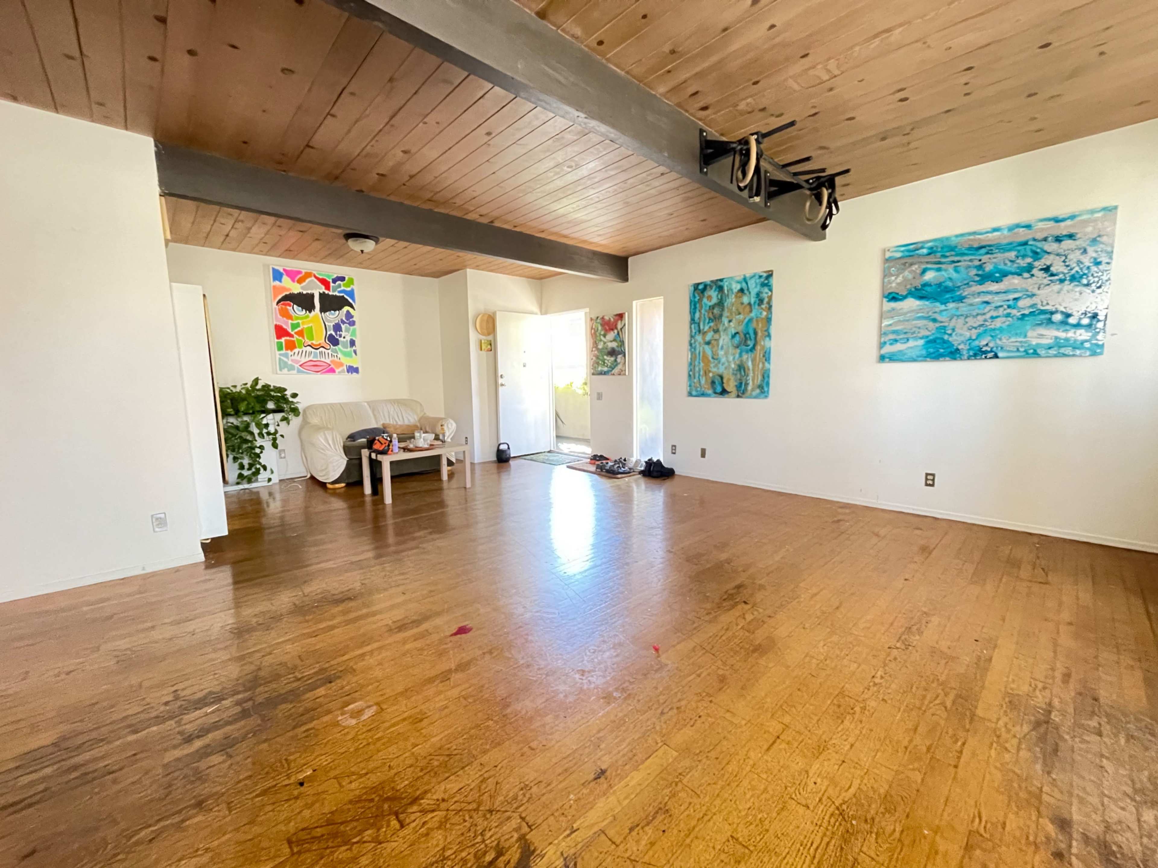 A spacious room features wooden flooring, light-colored walls, and various pieces of artwork hanging on the walls.