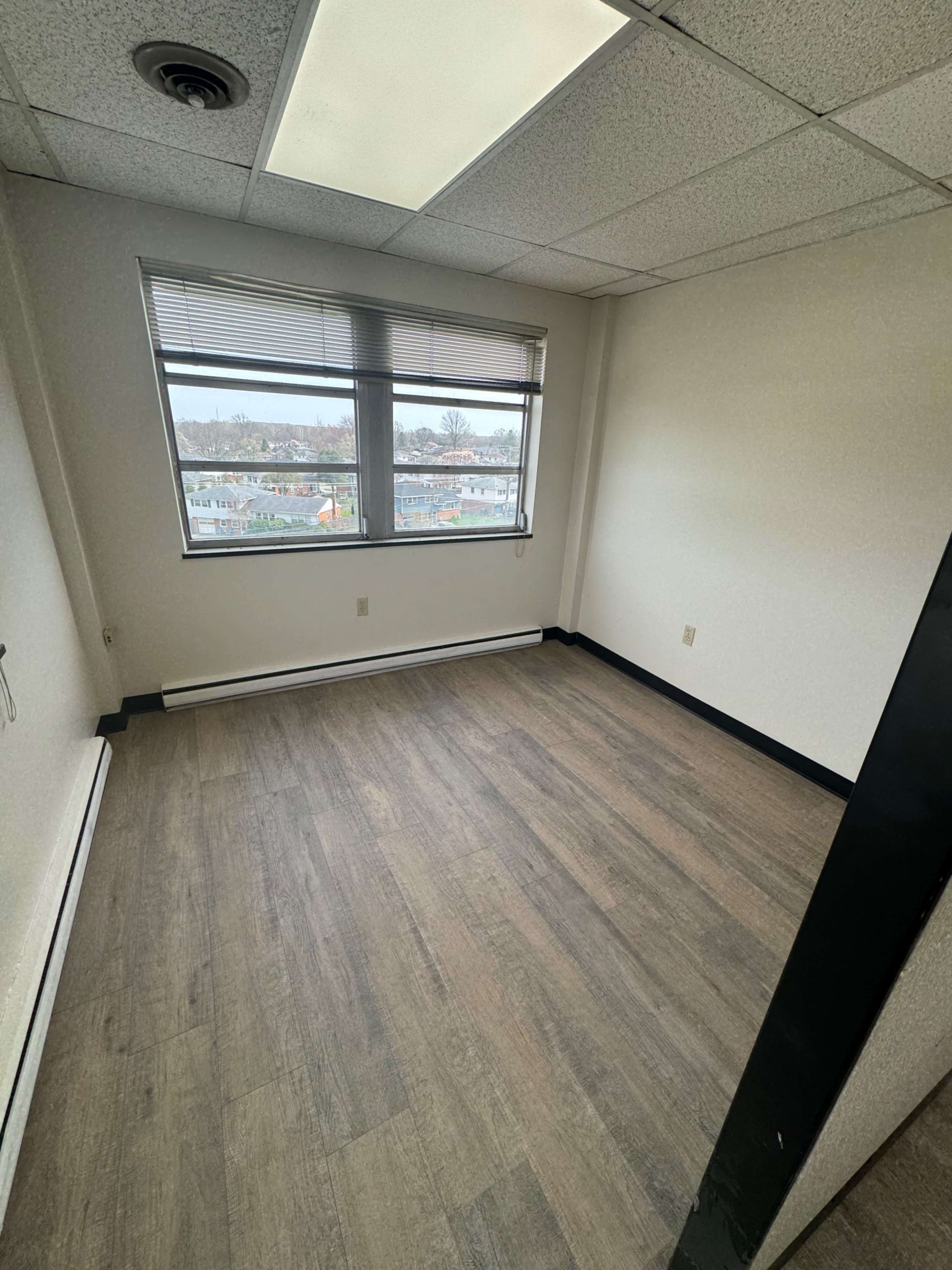 The image shows an empty office room with a large window and a wooden floor.