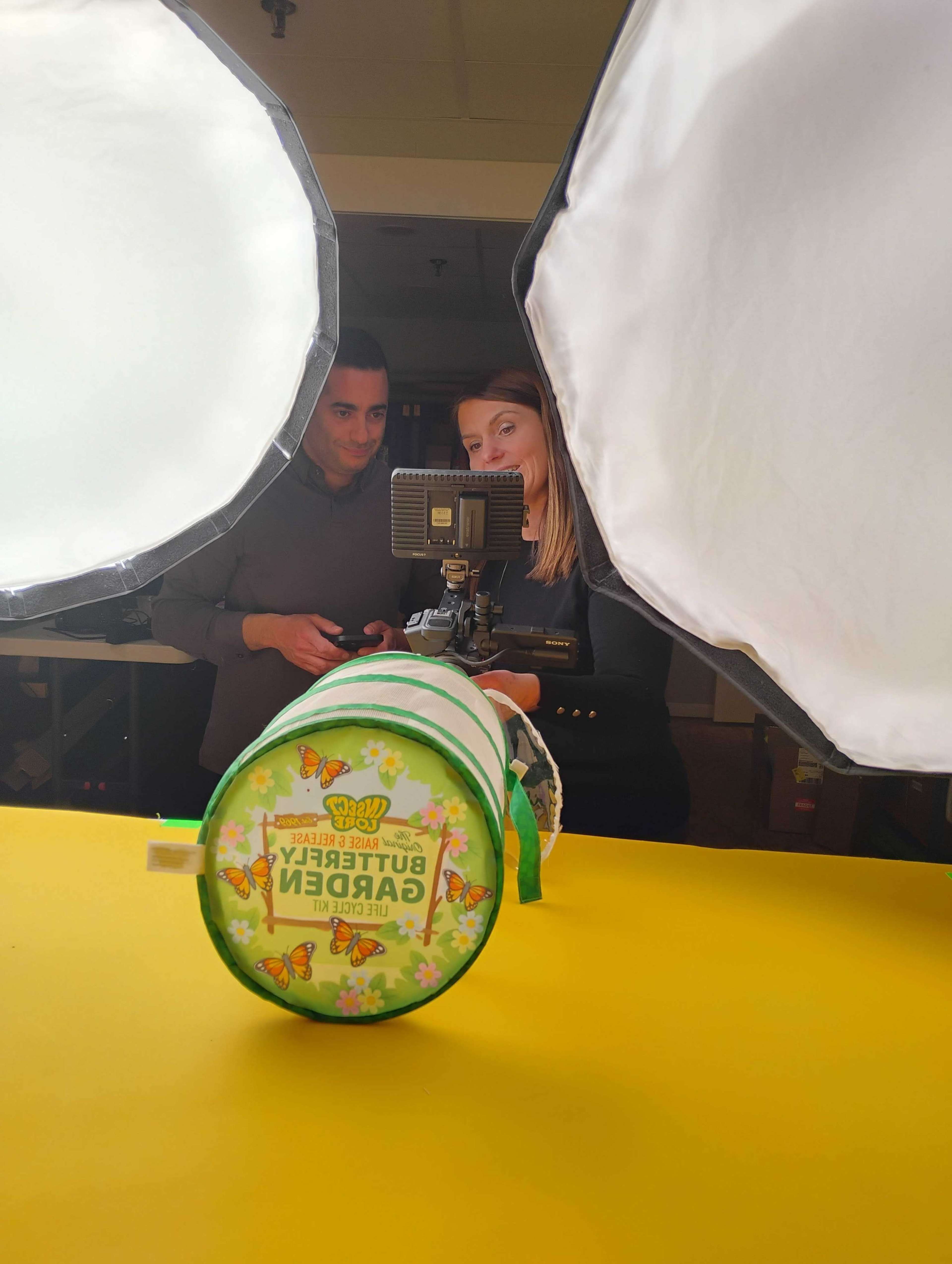 Two individuals stand behind a brightly colored display while setting up a camera with softbox lights.