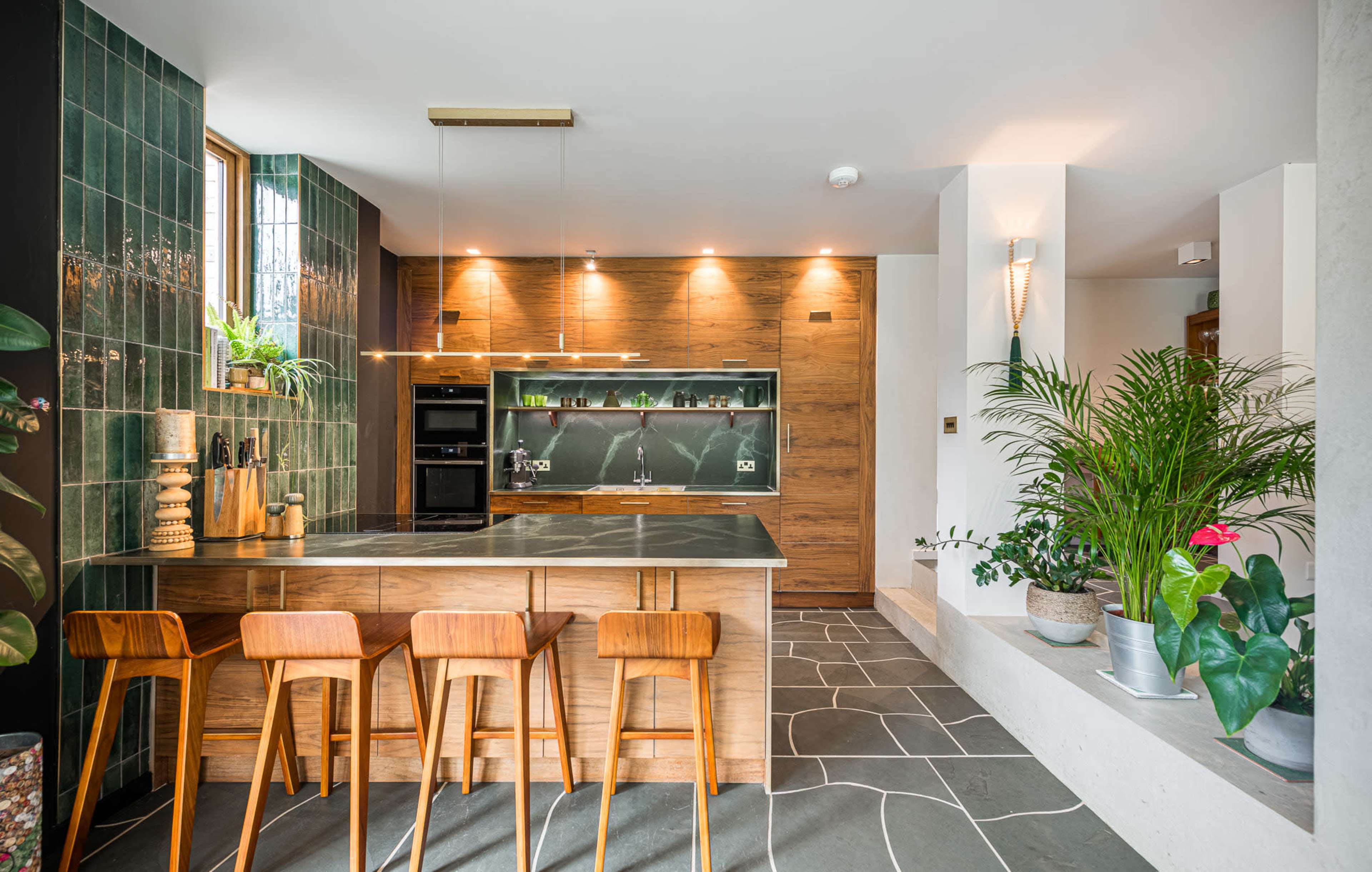 The image shows a modern kitchen with wooden cabinetry, a stone countertop, and green tiles, accompanied by potted plants and bar seating for four.