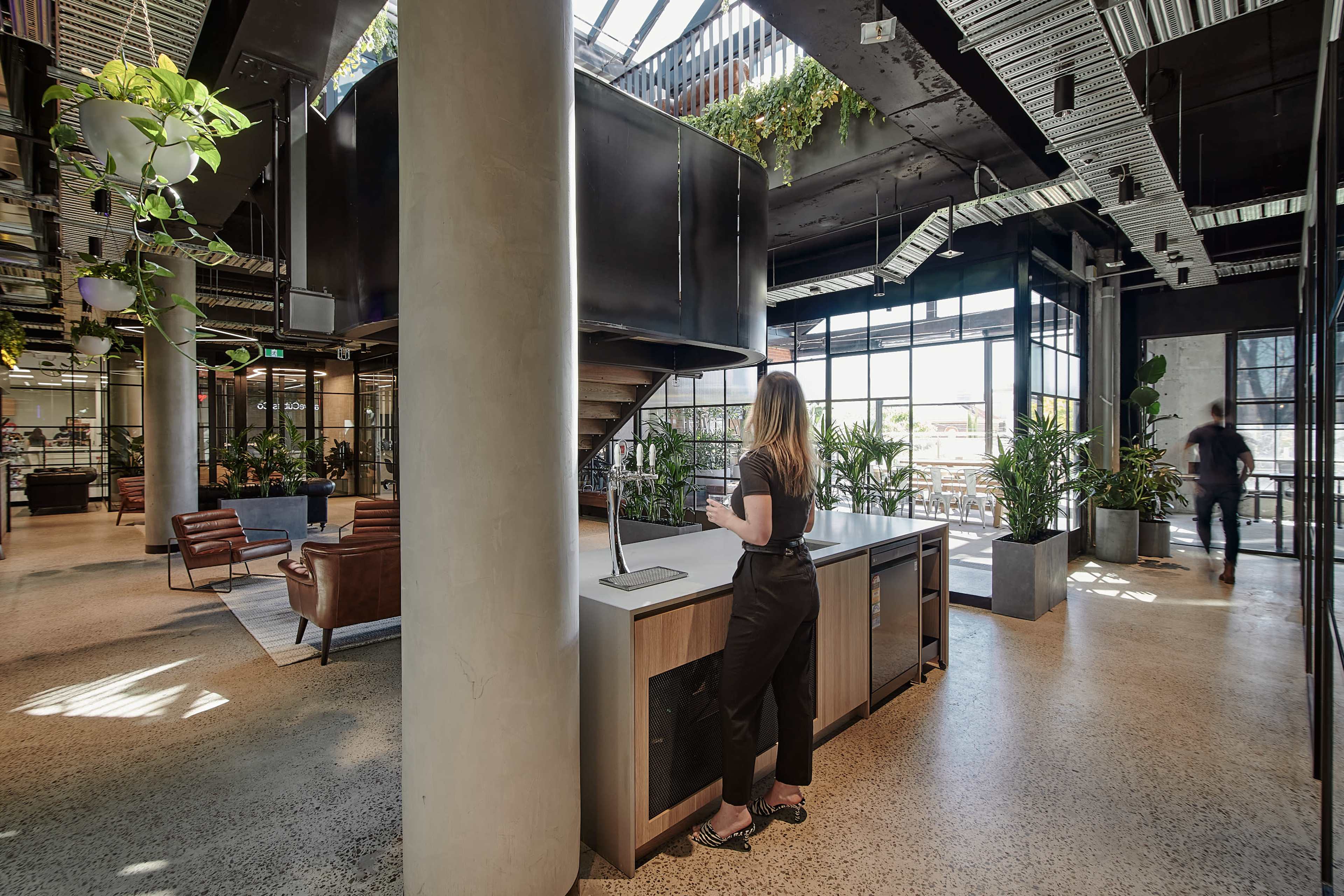 A woman stands at a kitchen island in a modern, open-concept office space filled with plants and large windows.