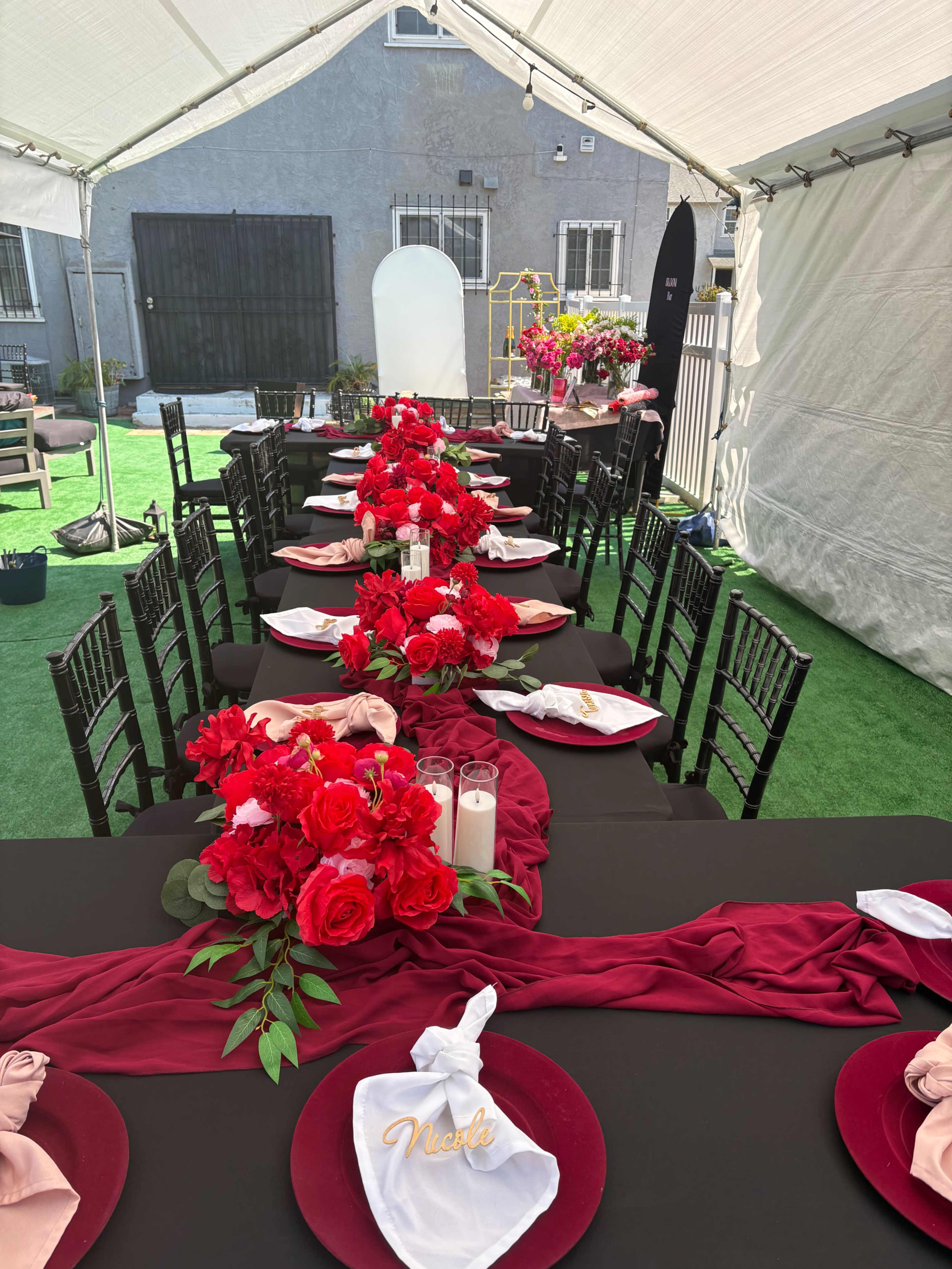 A long banquet table is set under a tent, adorned with red roses and white candles, featuring black tablecloths and decorative place settings.