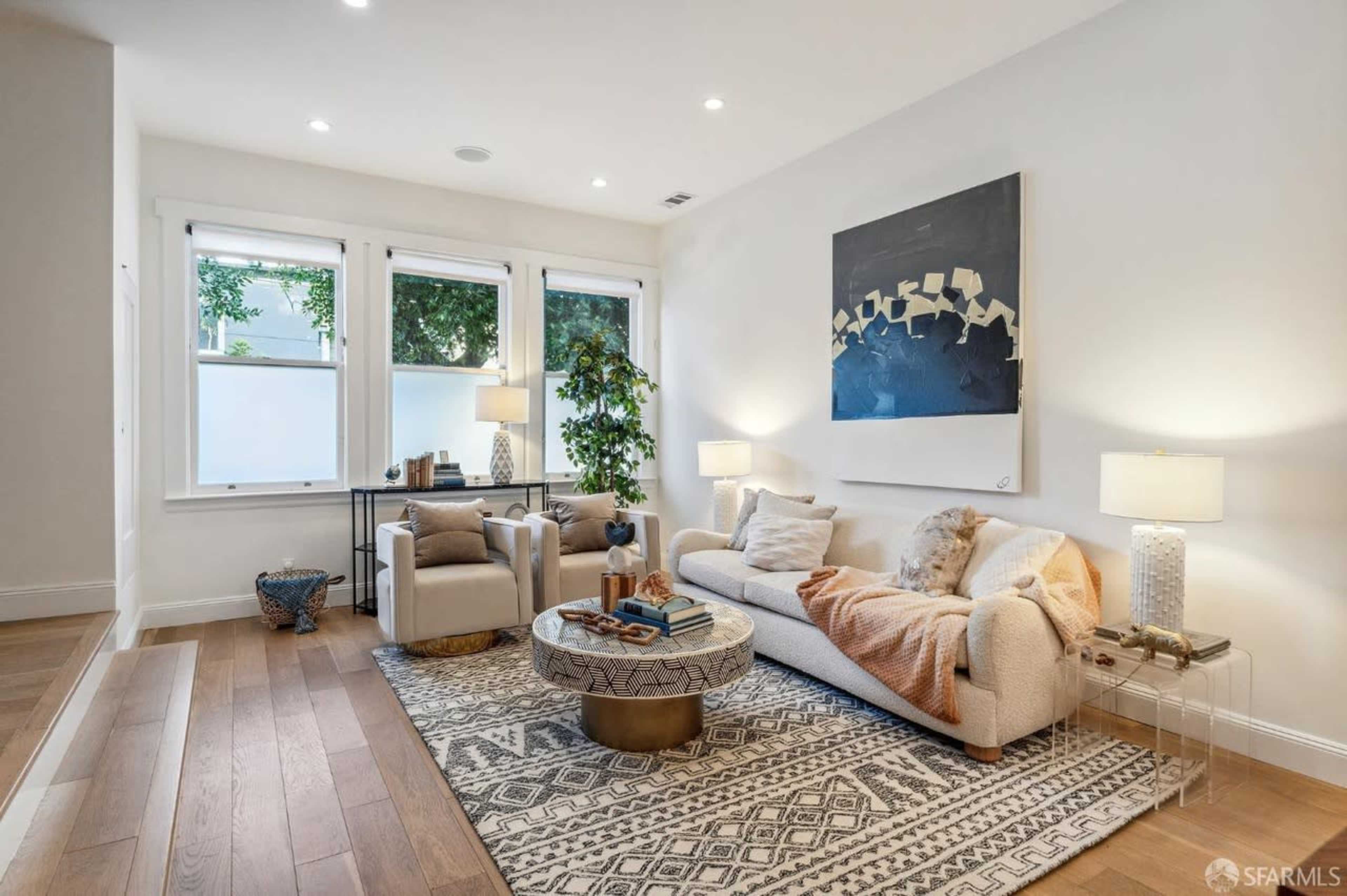 A well-lit living room featuring a beige sofa, two armchairs, a round coffee table, and large windows with greenery outside.