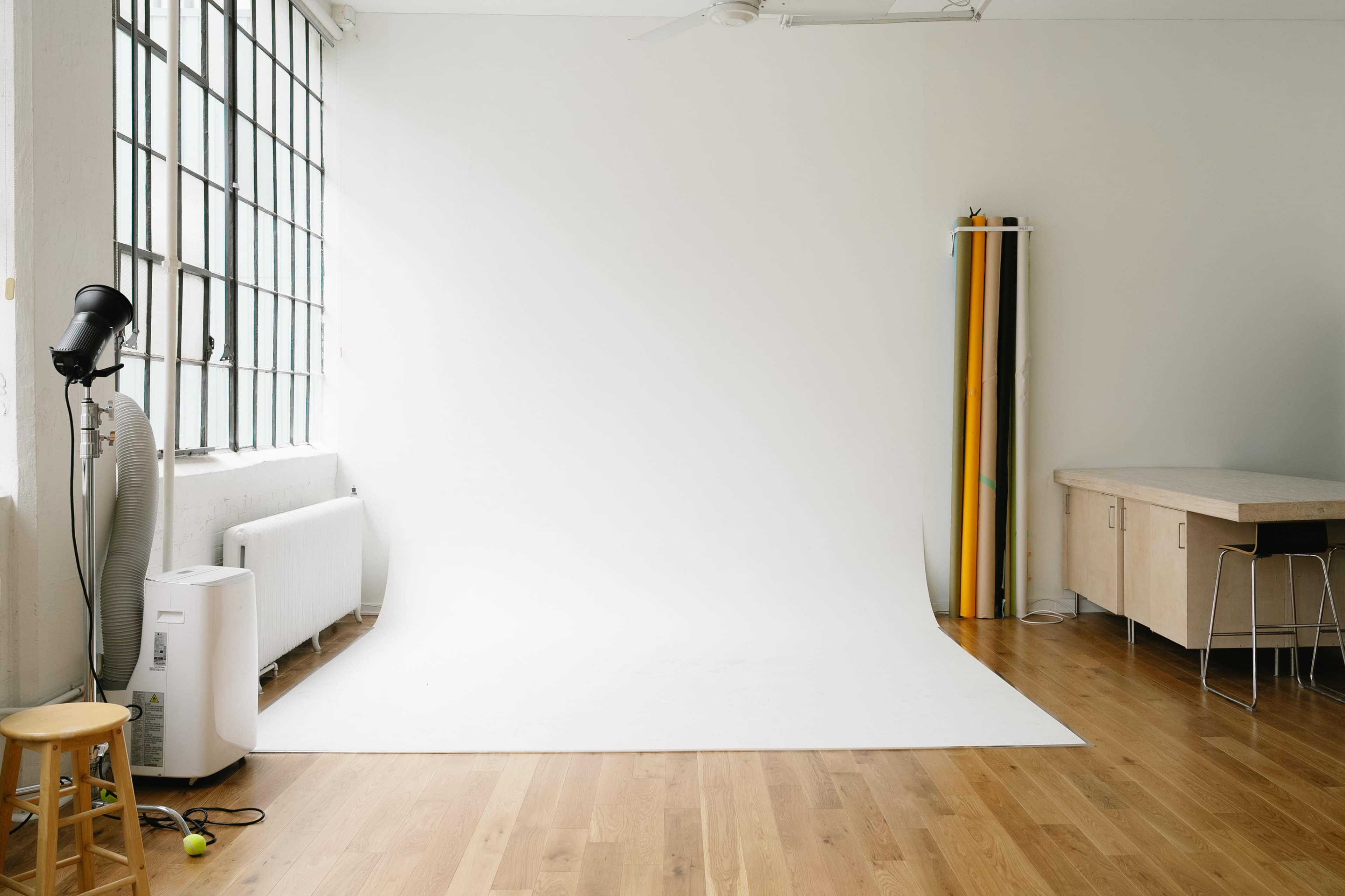 The image shows a bright, minimalistic studio space with a white backdrop, wooden flooring, and a stool.