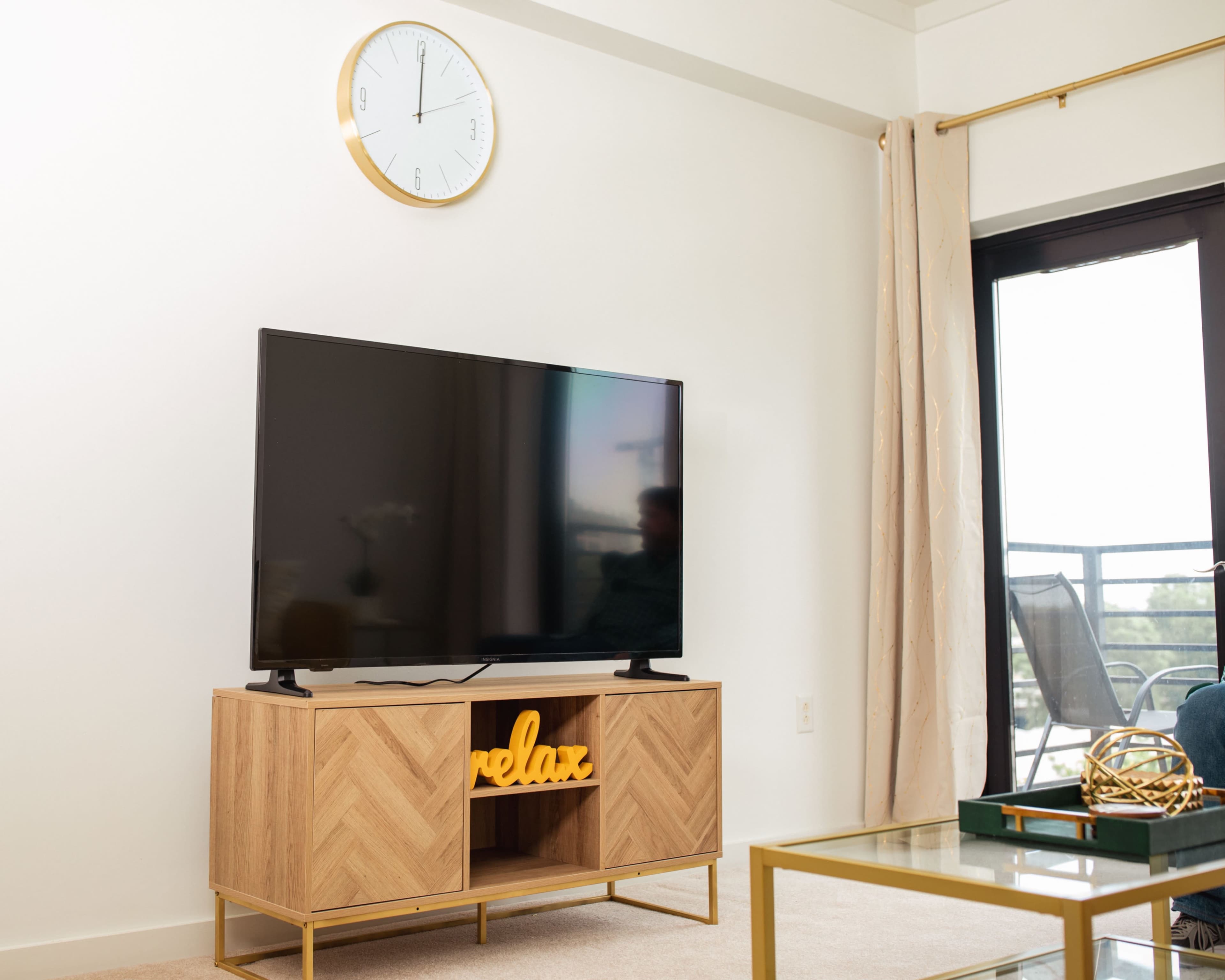 A modern living room features a television on a wooden stand with the word "relax" displayed, a wall clock, and a large window with a balcony view.