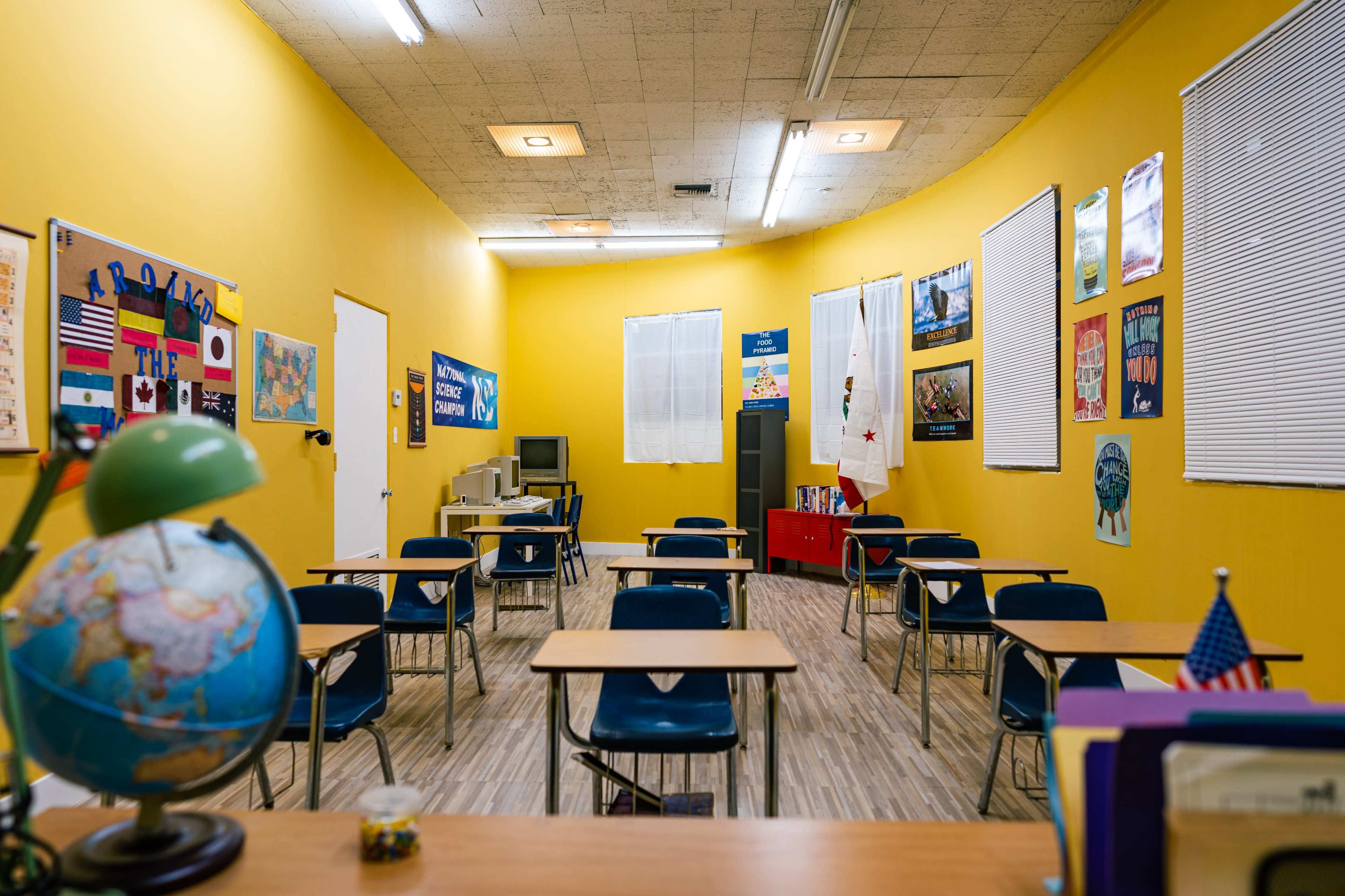 A classroom with yellow walls, empty desks arranged in a semicircle, and various educational posters on the walls.