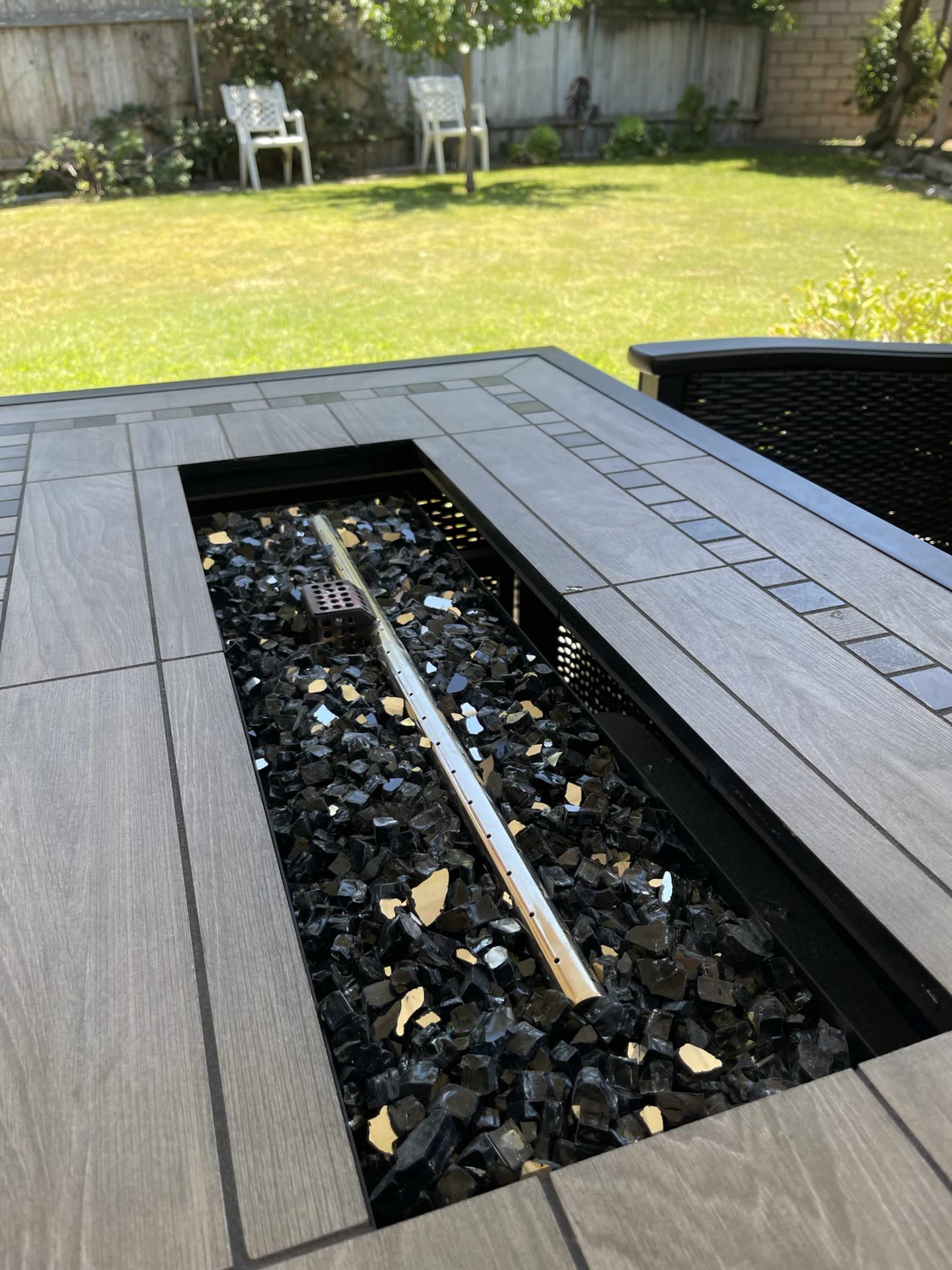 A patio table features a central fire pit filled with black and clear glass stones, surrounded by a grassy backyard and outdoor seating.