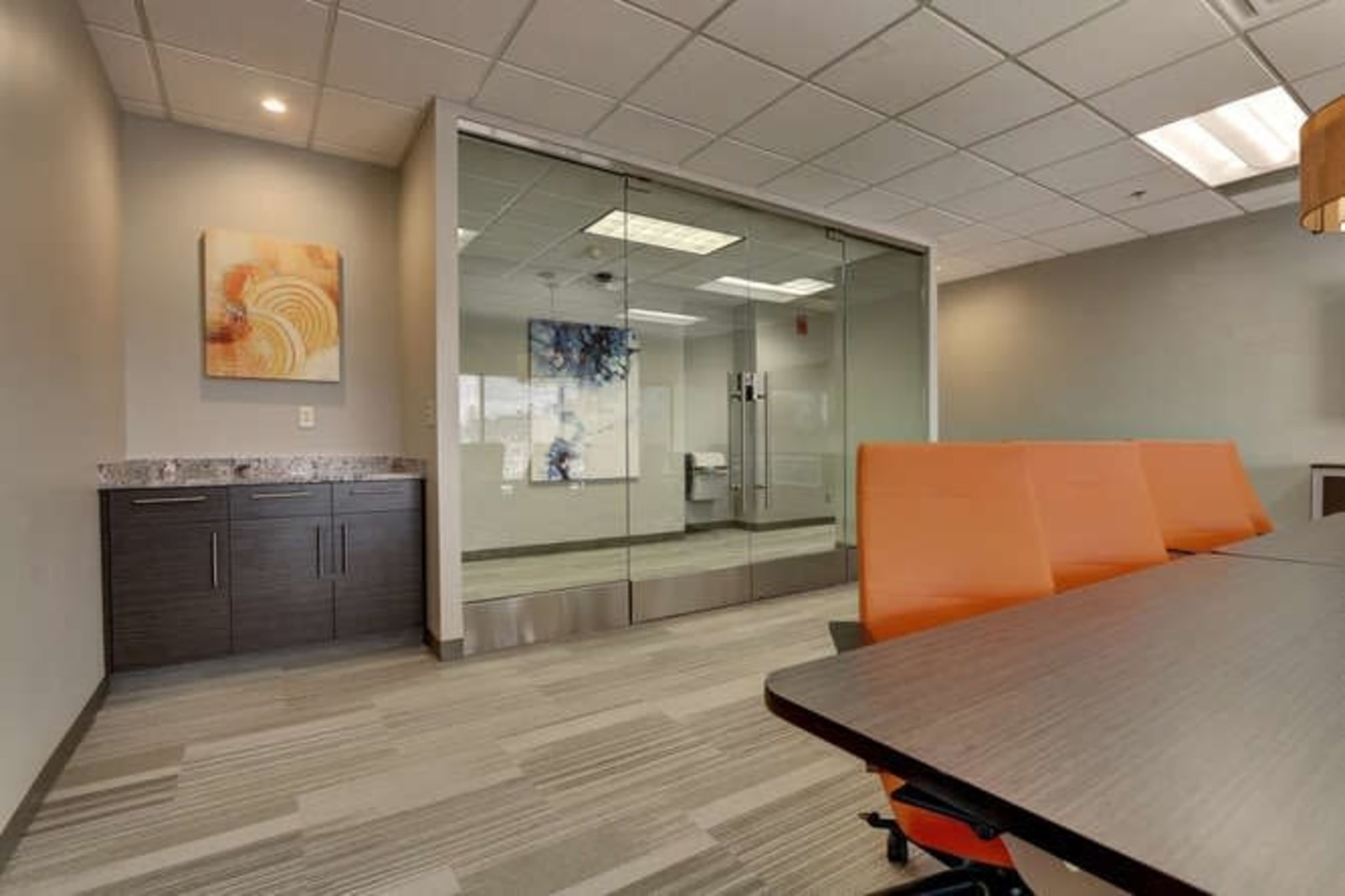 The image shows a modern office space featuring a conference table with orange chairs, a glass-walled meeting room, and a wall-mounted abstract artwork.