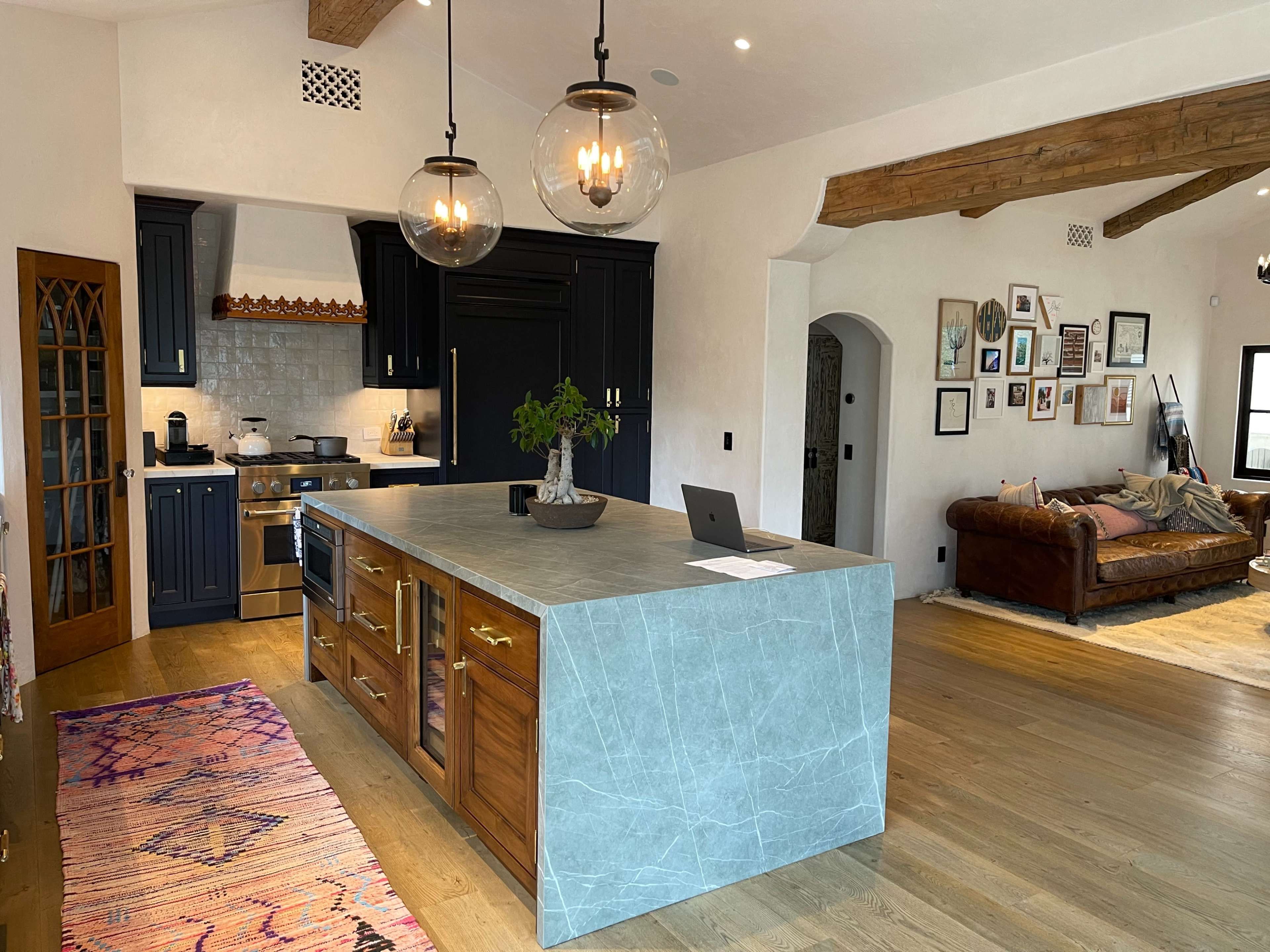 A modern kitchen with a large island featuring a mix of wooden and stone materials, surrounded by a living area with a brown leather sofa and framed pictures on the walls.