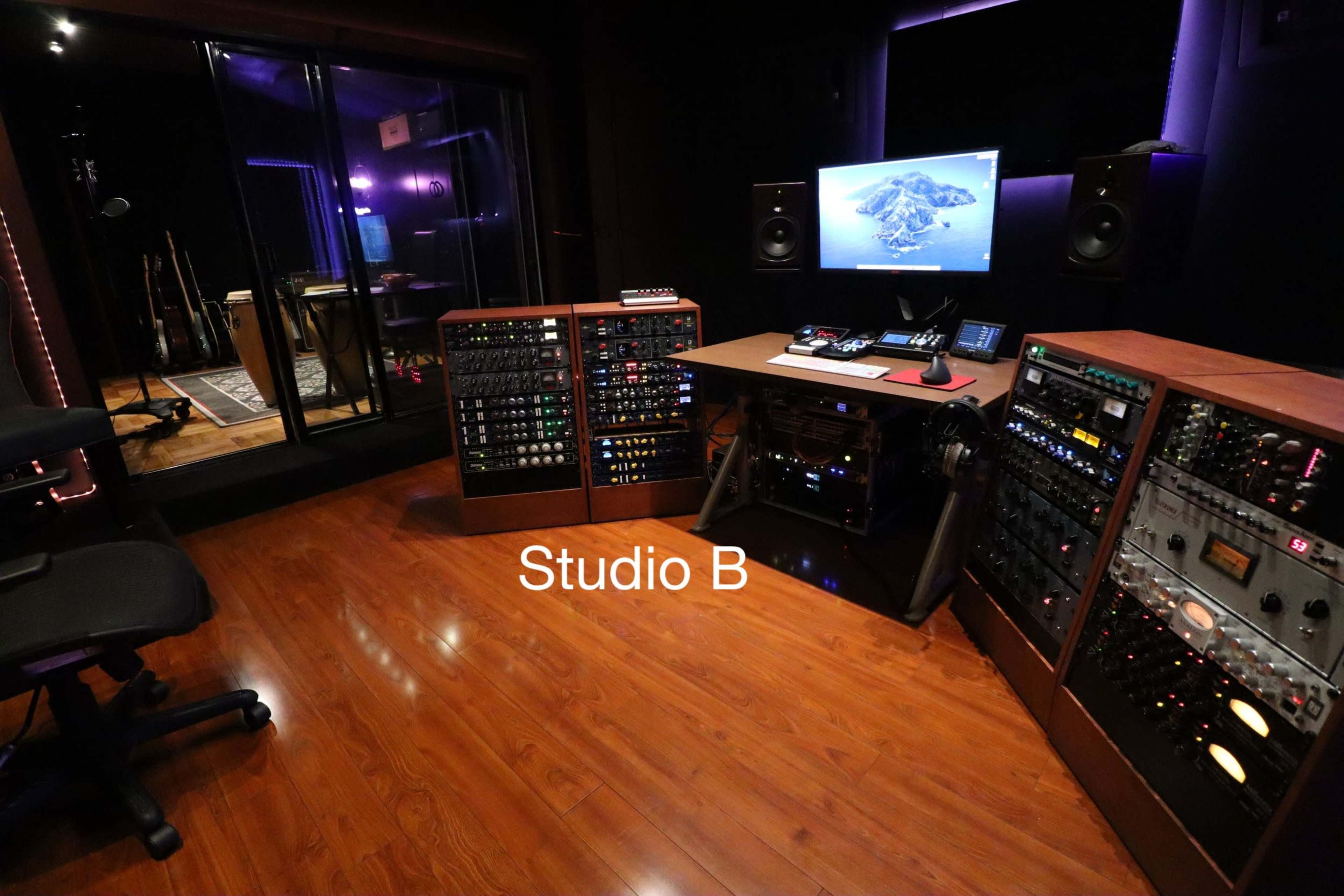 The image shows a music recording studio with various audio equipment, a large computer monitor, and a visible soundproof room in the background.