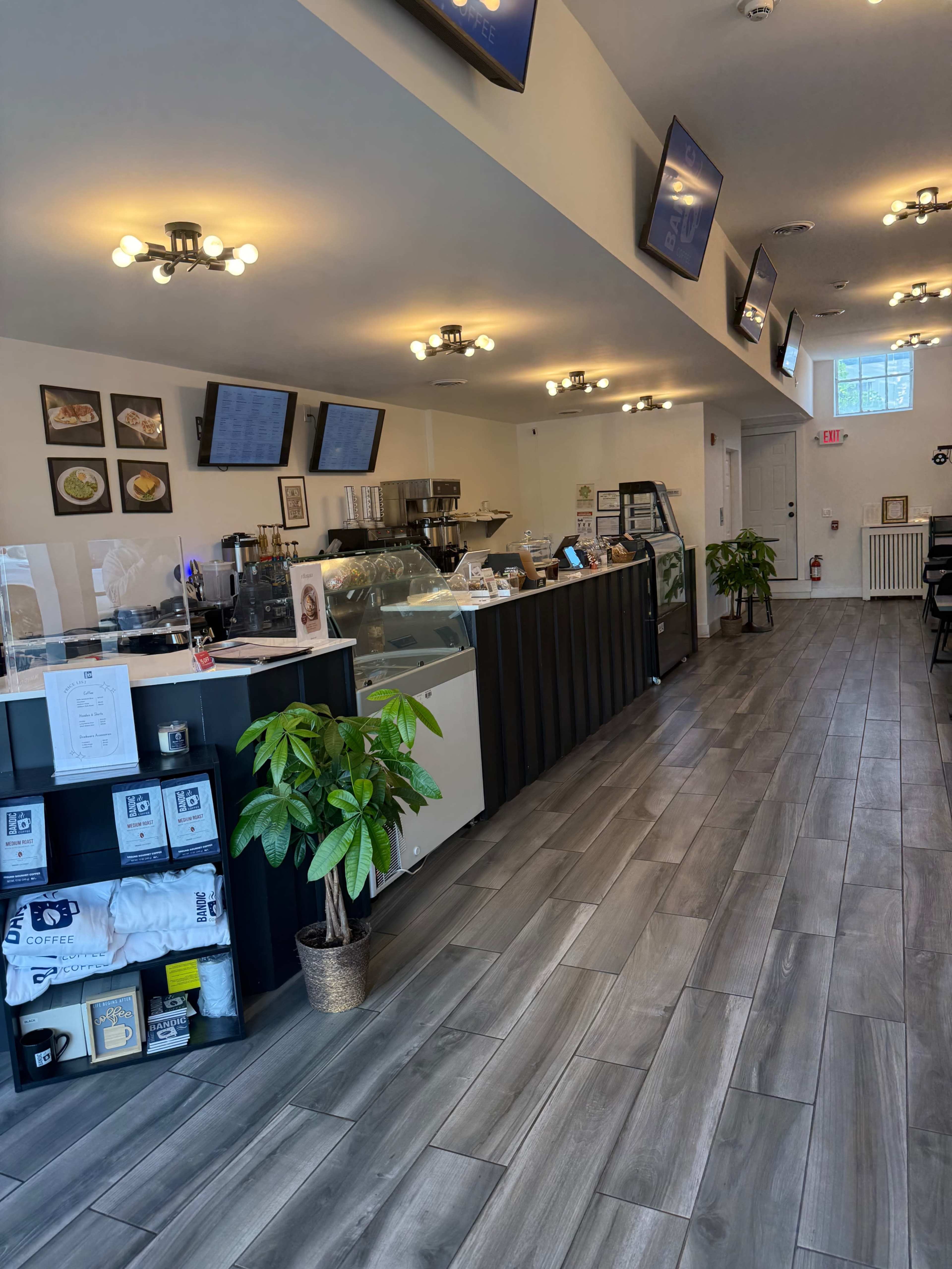 The image shows the interior of a modern café, featuring a counter with display cases, seating areas, and multiple screens mounted on the walls.