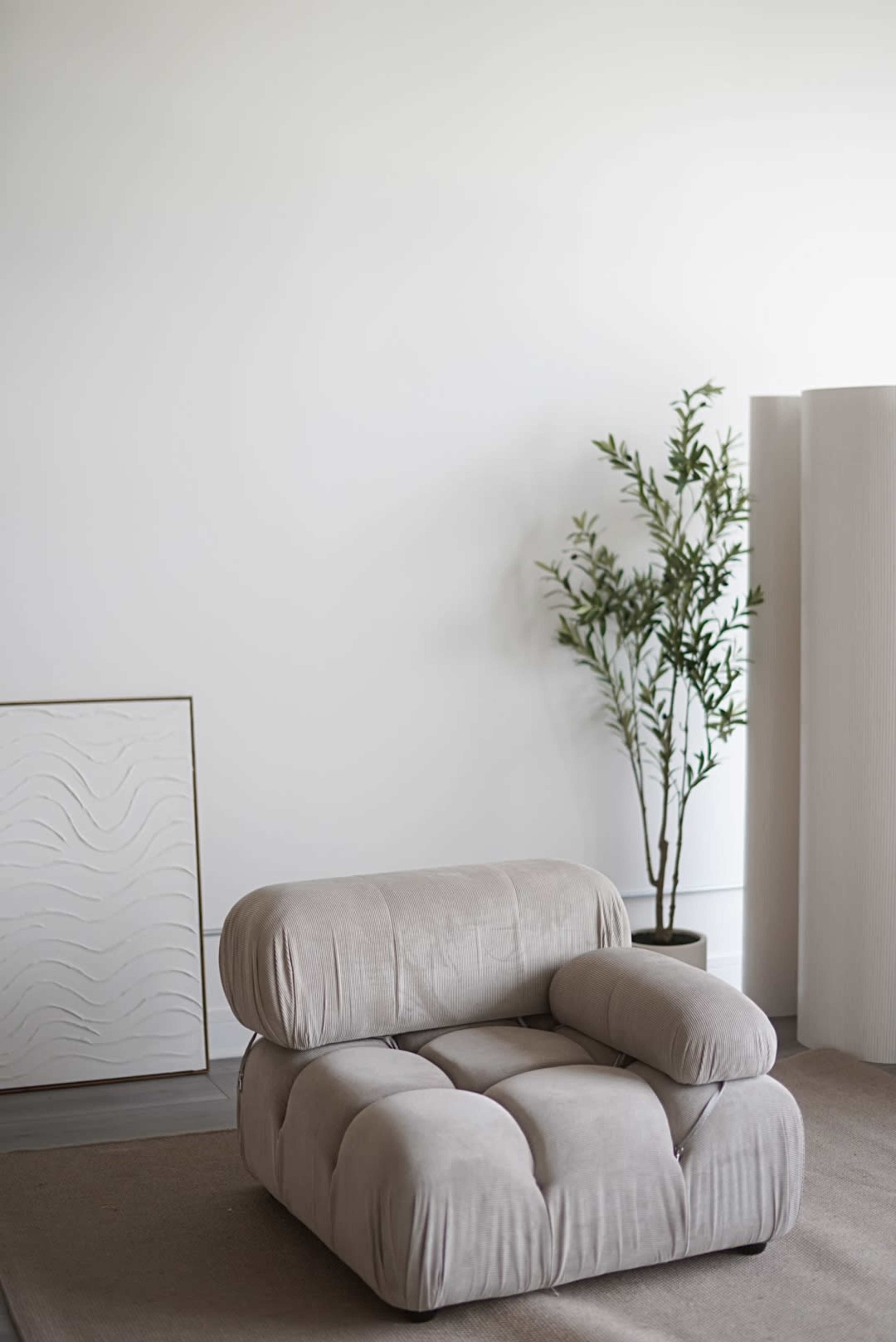 A plush, modern armchair in a neutral color sits on a carpet in a minimalist room, accompanied by a small potted plant and a textured wall decoration.