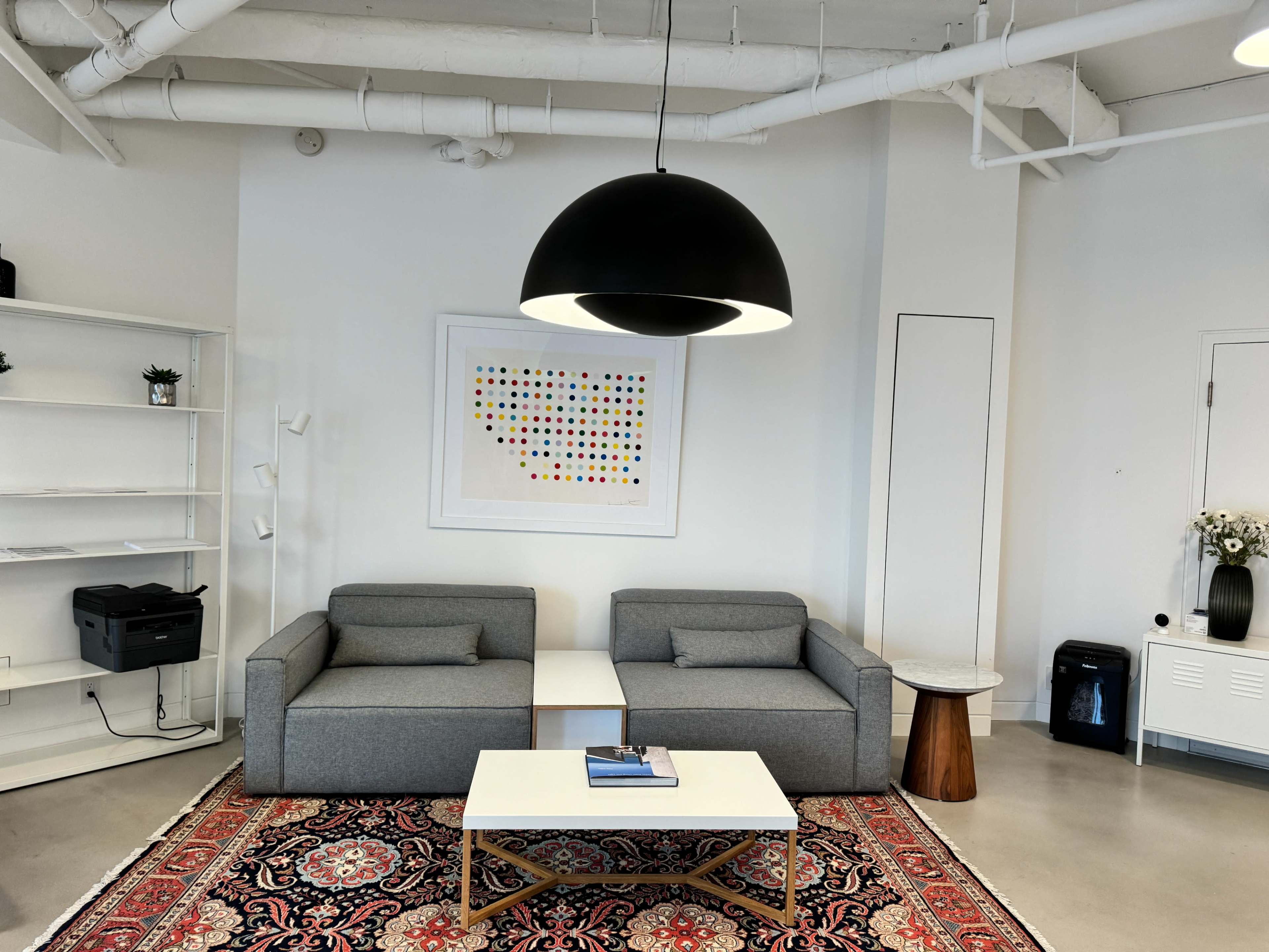 A minimalist lounge area features two gray sofas, a round black pendant light, and a decorative rug, with a wall art piece and shelving in the background.