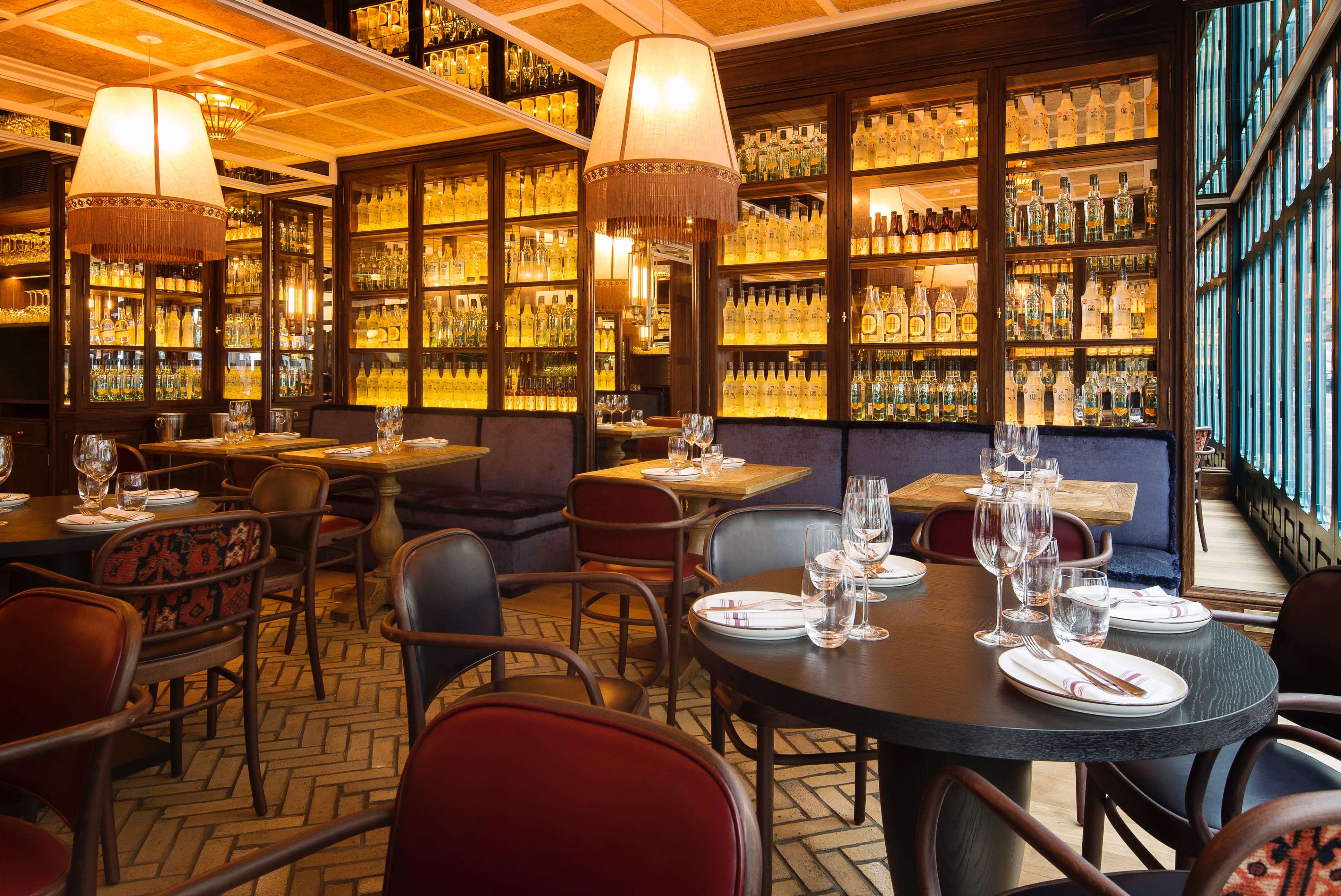 The image shows a warmly lit restaurant interior featuring elegantly set tables, wooden shelves filled with bottles, and plush seating arrangements.