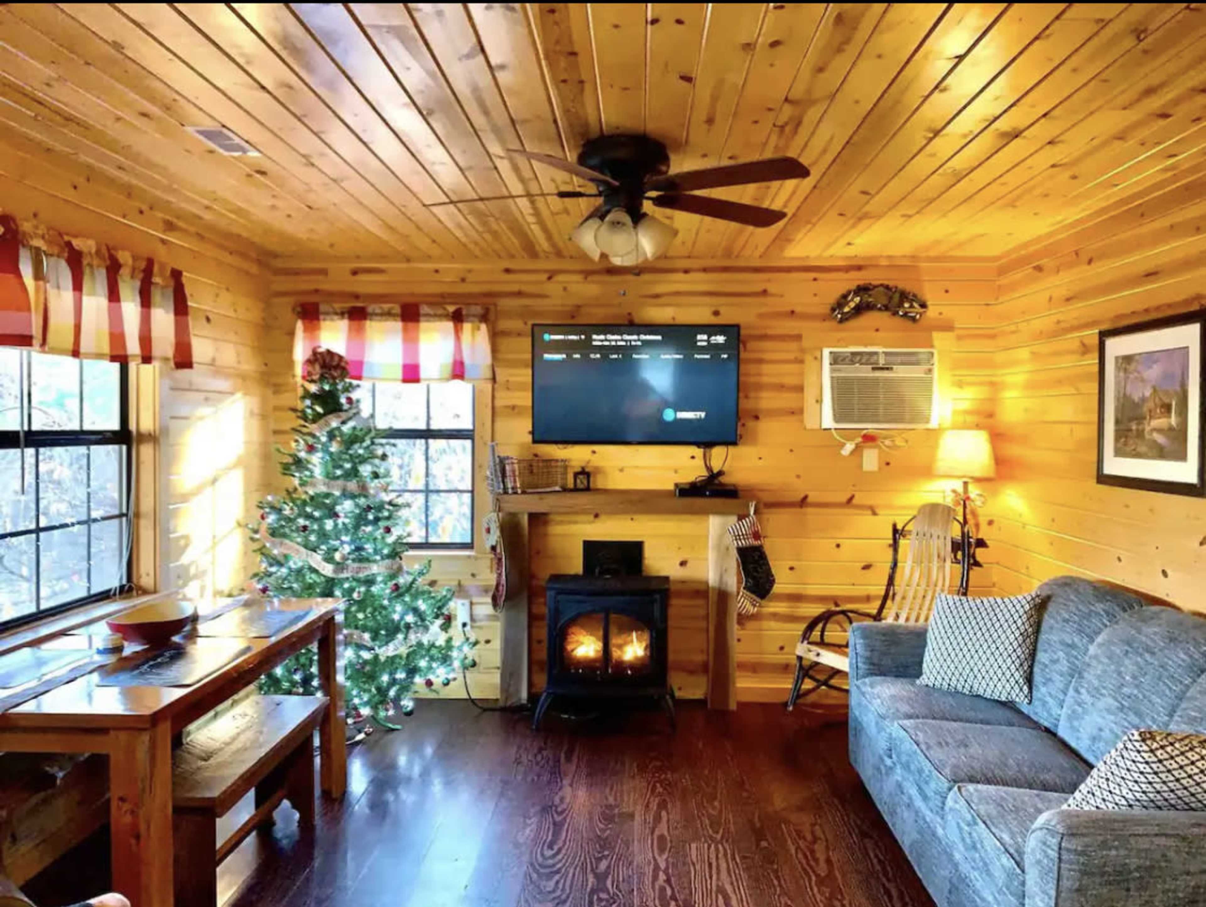 A cozy living room features wooden walls, a Christmas tree, a fireplace, and a couch.