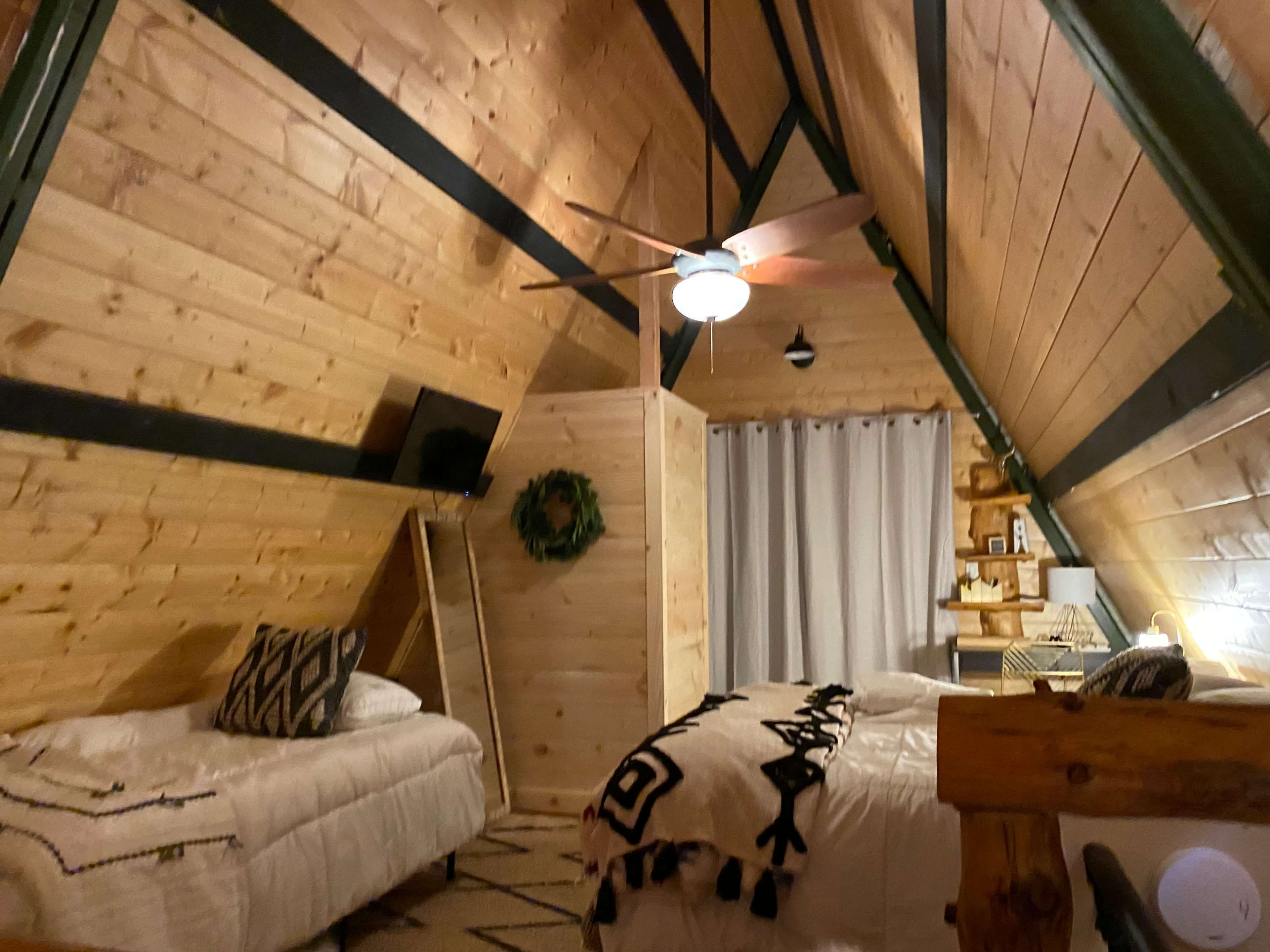 A cozy A-frame bedroom with wooden walls, a ceiling fan, a bed with white bedding, and a small separation for the closet area.