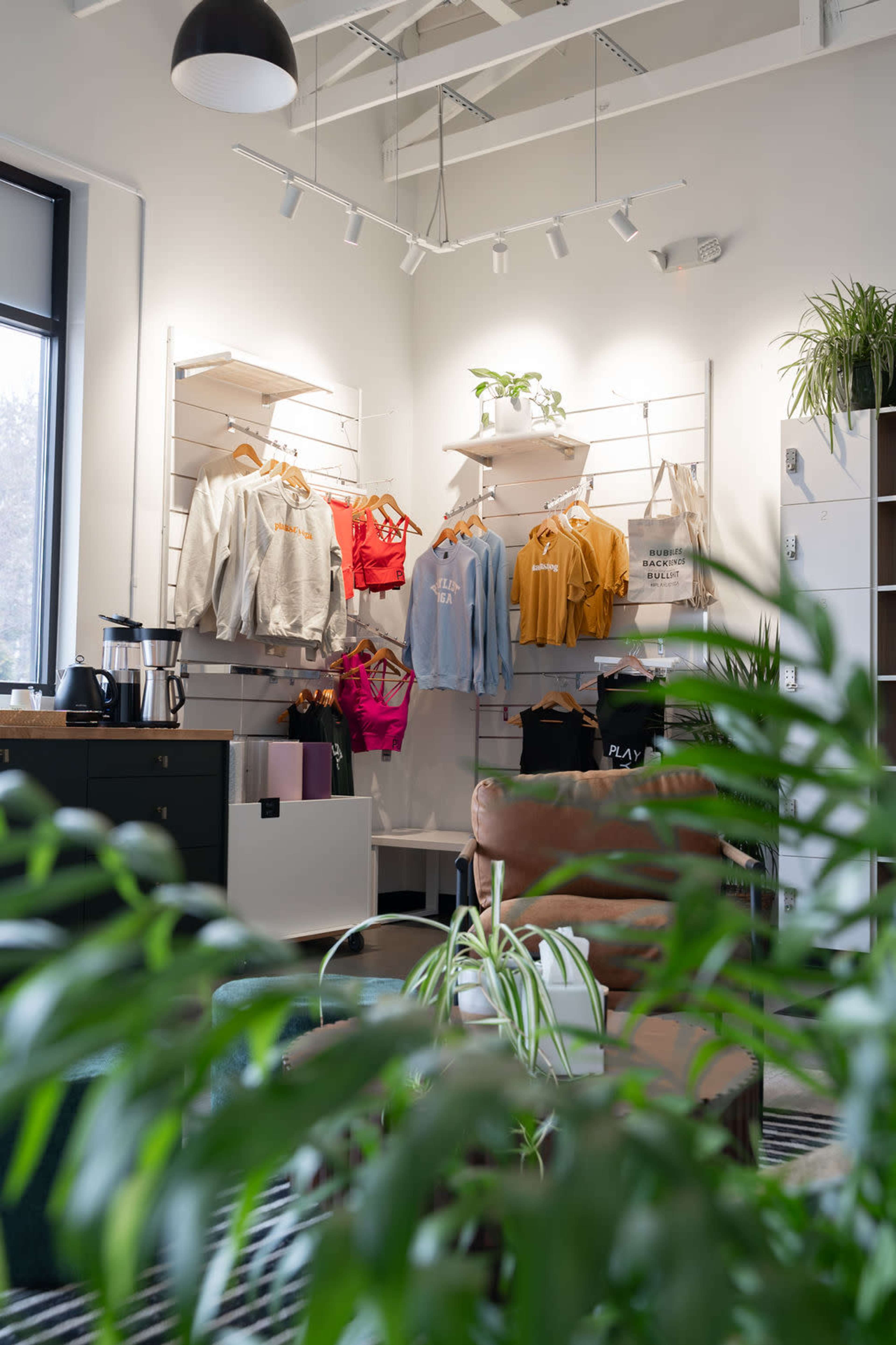 The image shows a modern retail space featuring a clothing display with various sweatshirts and hoodies hung on wall racks, alongside a cozy seating area and greenery.