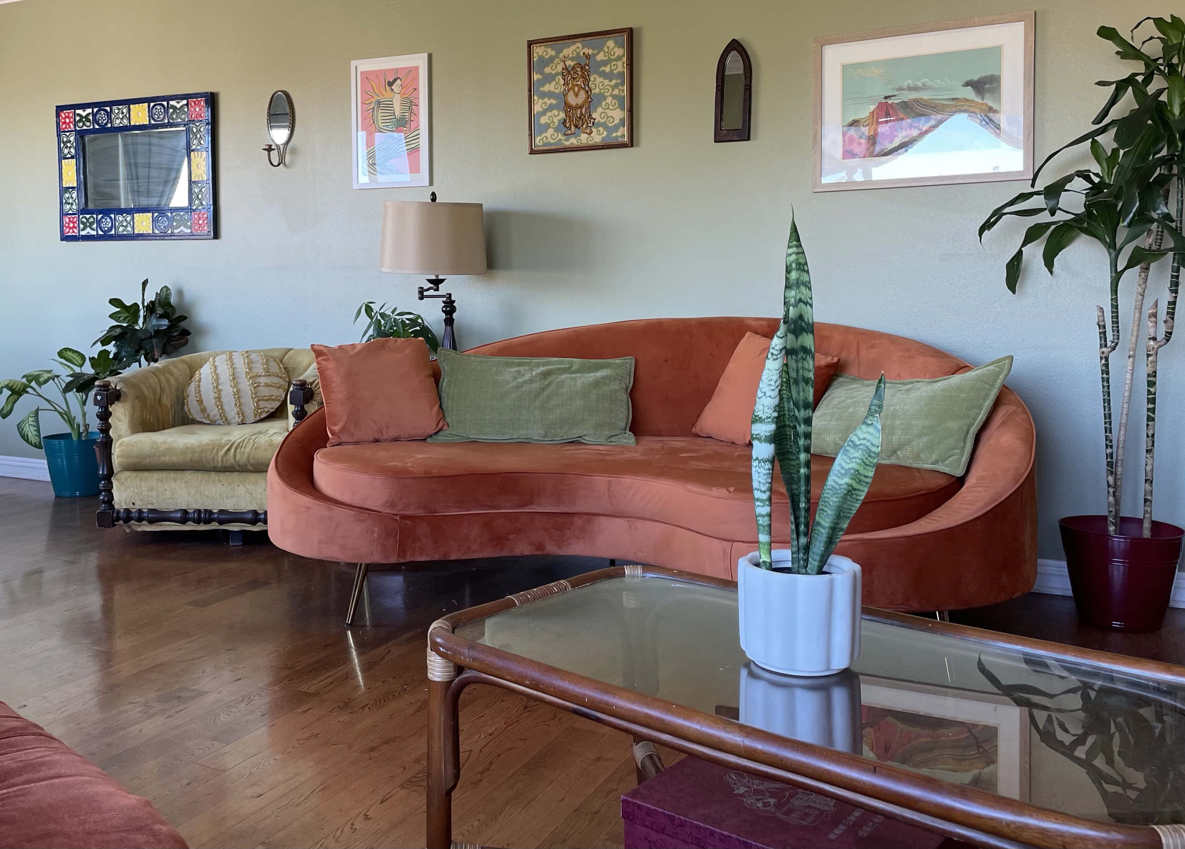 The room features an orange curved couch, a yellow armchair, a coffee table, and several plants against a green wall adorned with various framed artworks.