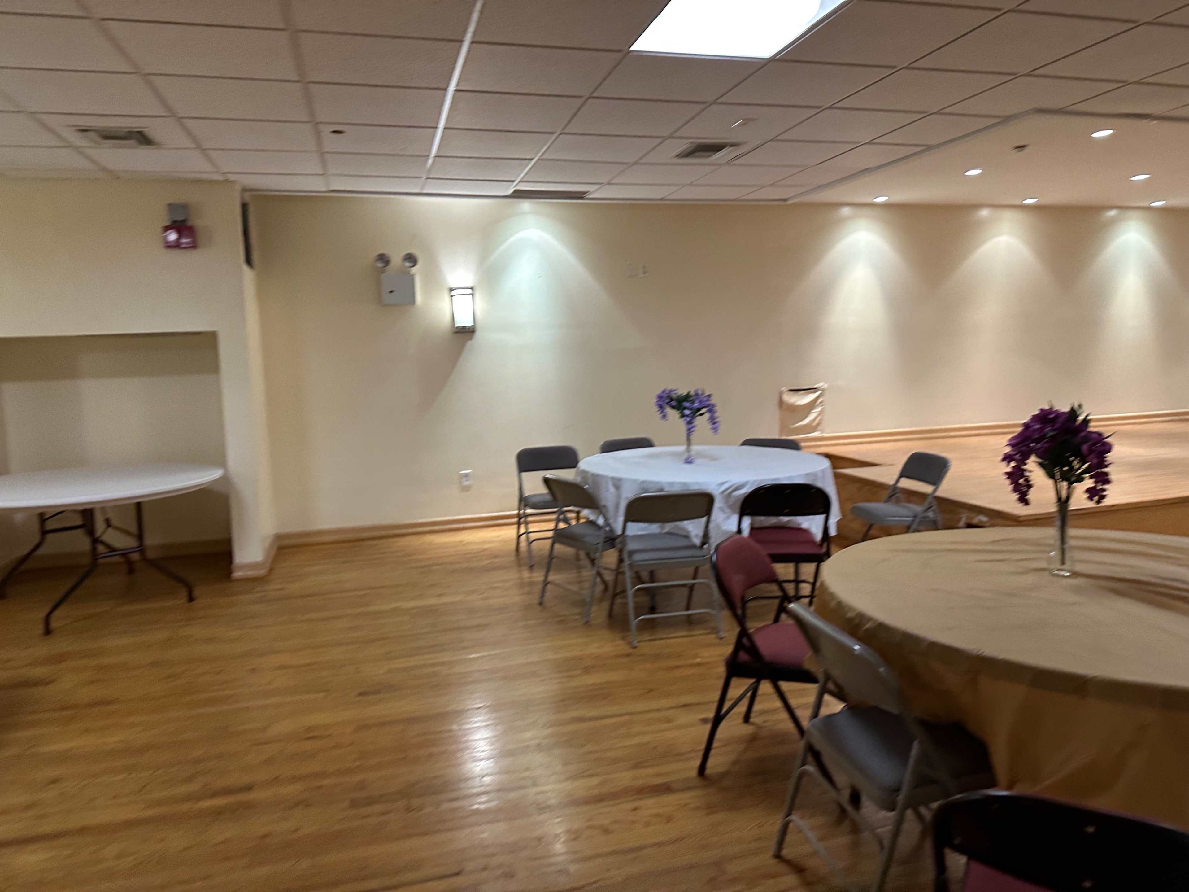 A spacious room with wooden floors contains several tables covered with tablecloths and chairs arranged around them, with a small stage at the front.