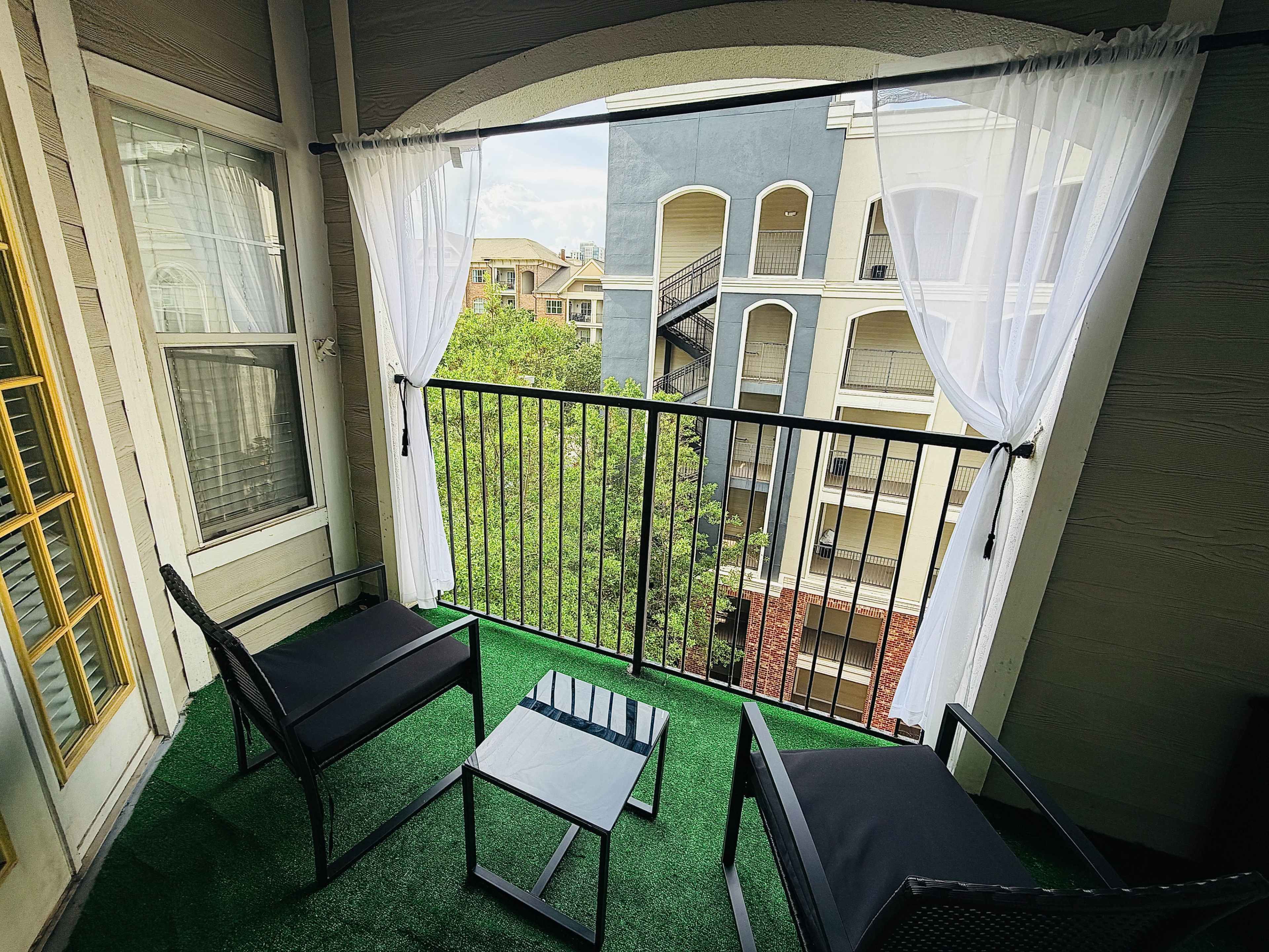A small balcony features two black chairs and a glass table, with sheer white curtains framing the view of neighboring buildings.