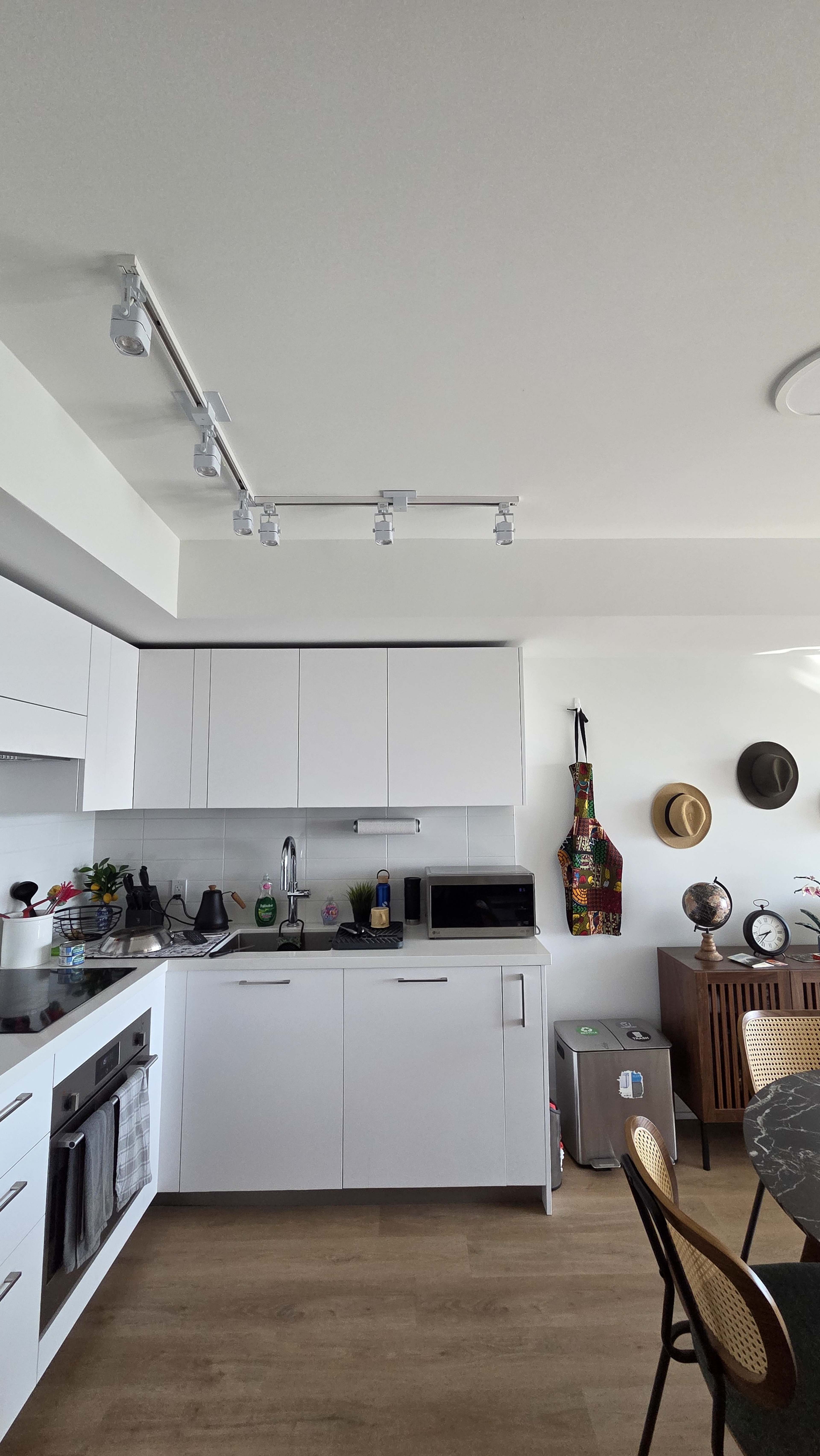 The image depicts a modern kitchen with white cabinets, a countertop, and various appliances, including a microwave and a refrigerator, alongside a dining table and decorative items on the walls.