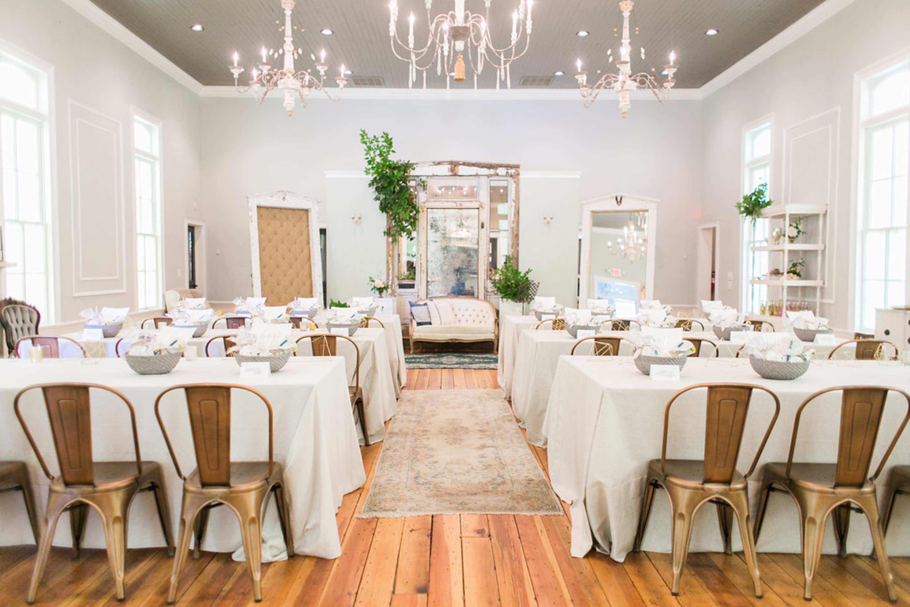 A spacious event venue with several tables dressed in light linens, arranged neatly around a central area featuring a large mirror and decorative plants.