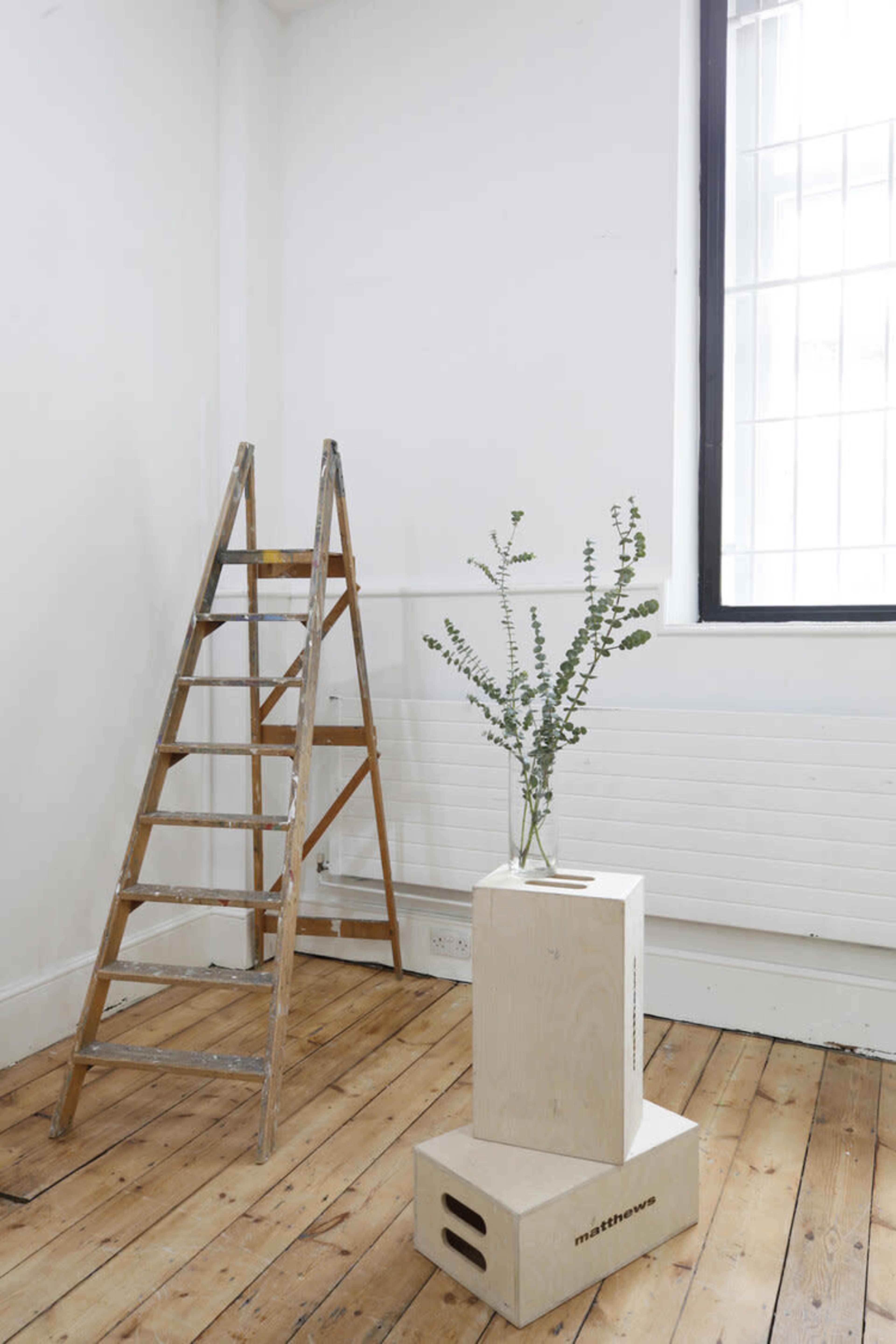 A wooden ladder stands next to a minimalist wooden box with a small vase containing greenery in a bright, sparsely decorated room.