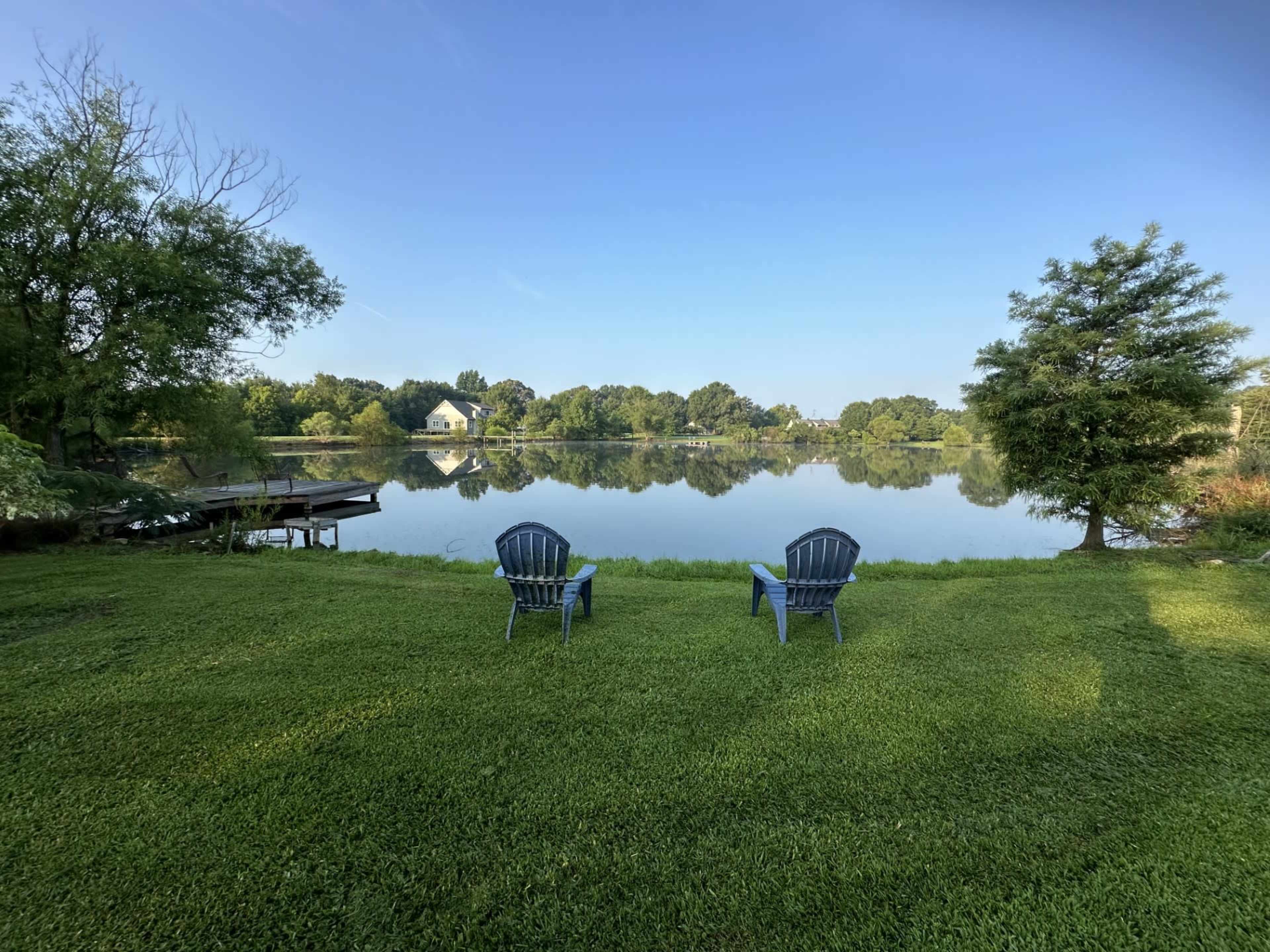 Two blue chairs sit on a grassy area by a calm lake, surrounded by trees and a clear blue sky.