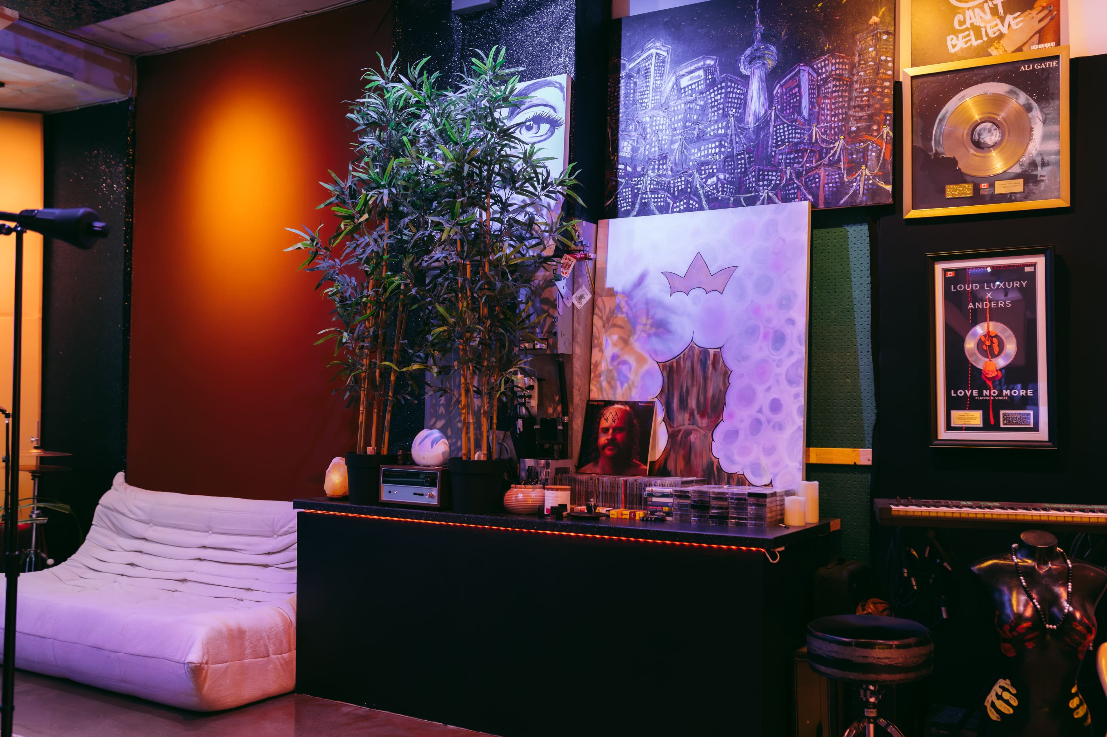 The image features a modern music studio corner with a white sofa, decorated walls showcasing artwork and framed records, and a potted plant.