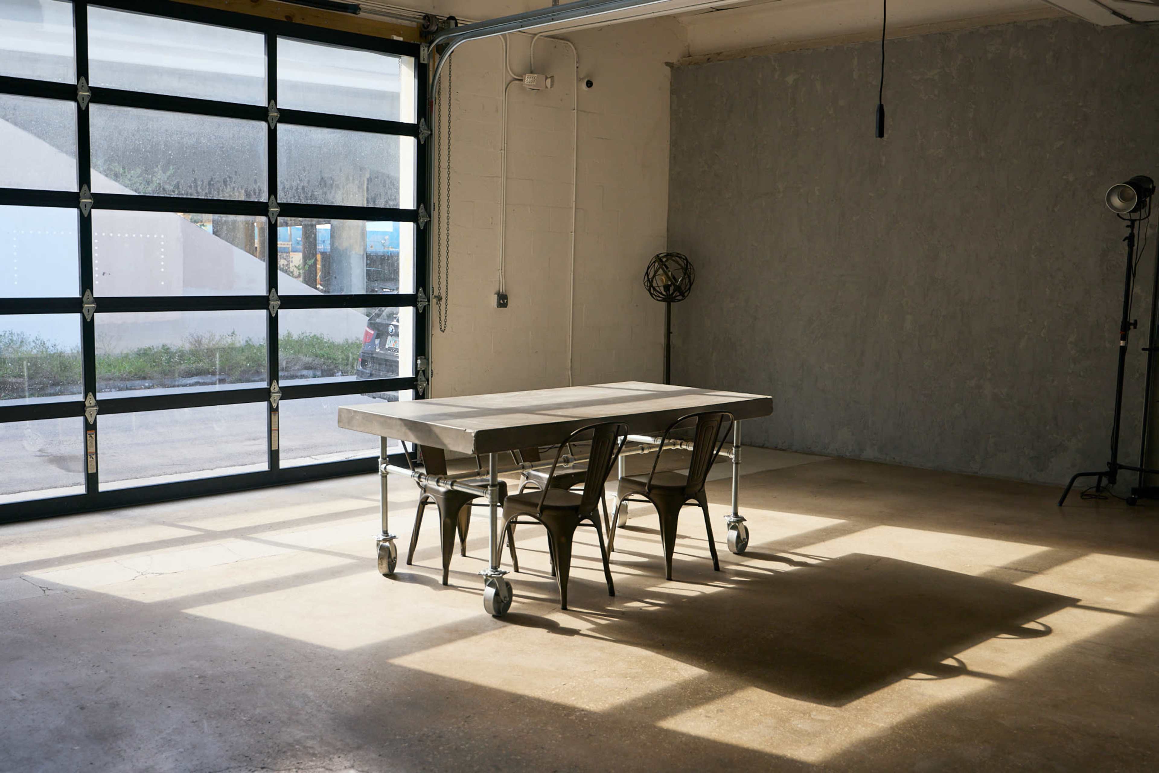The image shows a minimalist industrial space with a metal table on wheels and four matching chairs, illuminated by natural light from a large garage door.